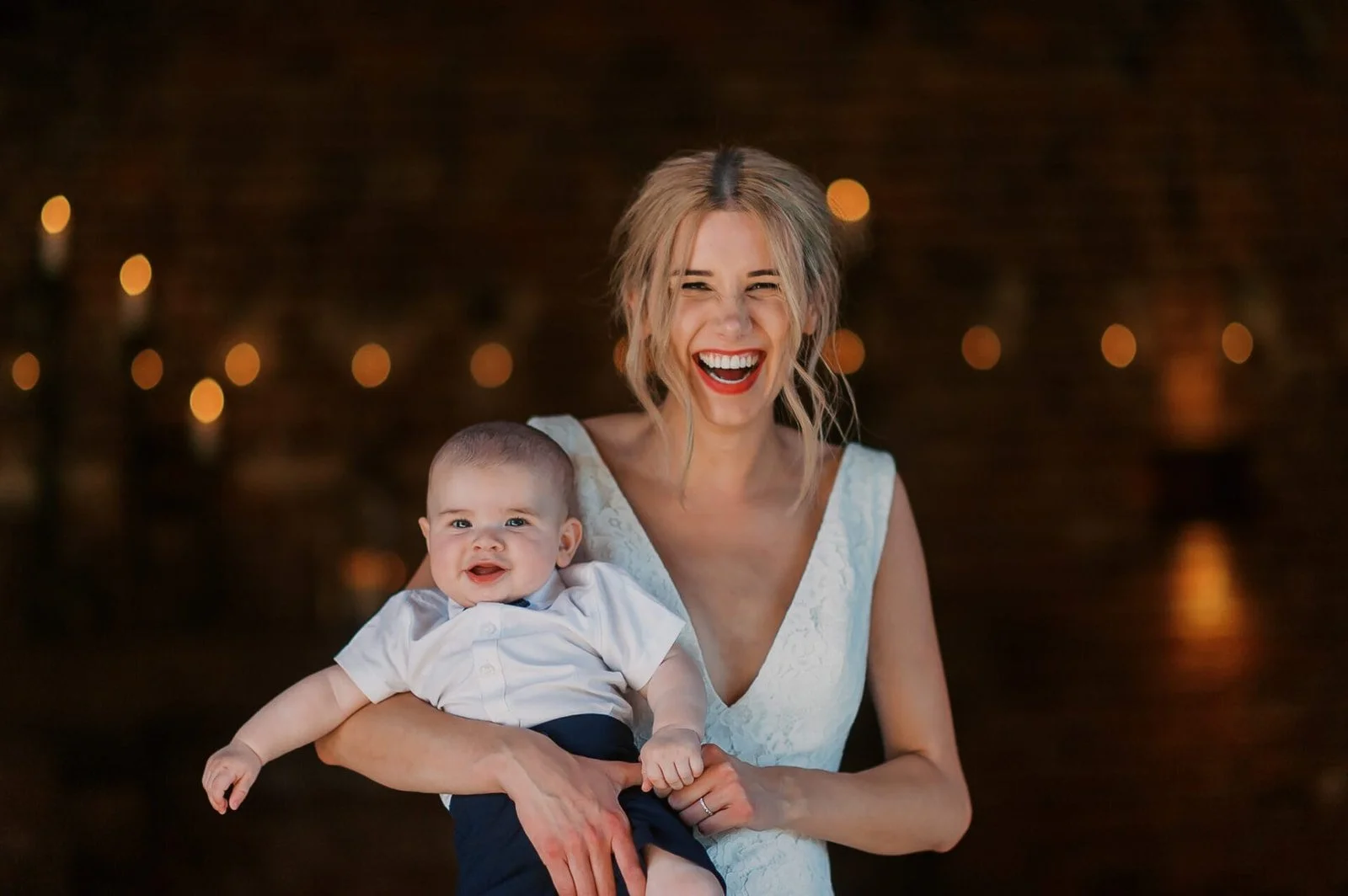 A woman in a white dress joyfully holds a smiling baby dressed in a white shirt and dark pants. The background is softly lit with warm, blurred lights, creating a cozy atmosphere.