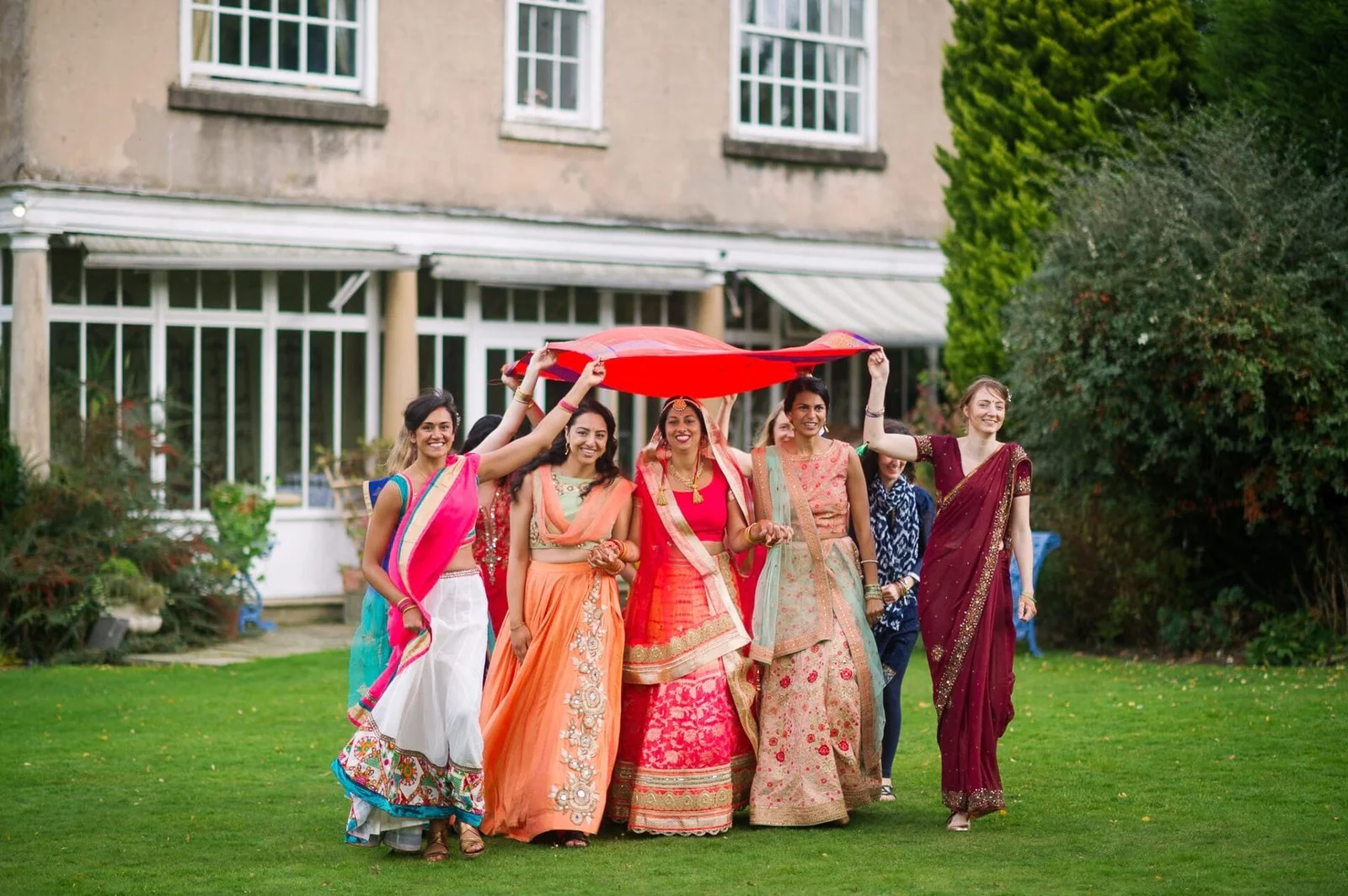 A group of smiling women in colorful traditional outfits walk across a lawn, holding a red canopy. They appear joyful and are positioned in front of a building with large windows.