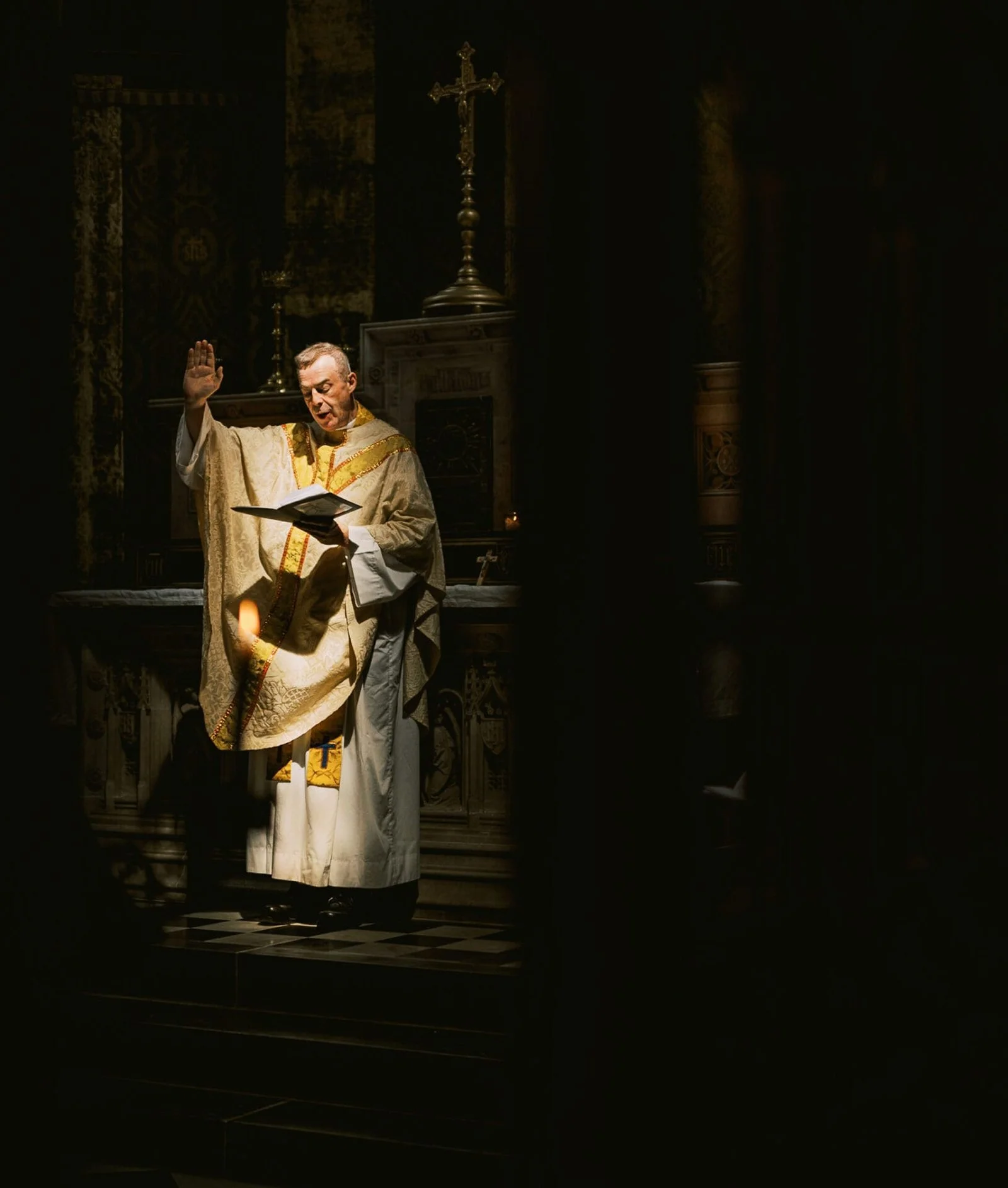 A priest in ornate robes stands near an altar, raising one hand in blessing while holding a book in the other. Soft, dramatic lighting highlights him against a dark background, with a cross visible behind.