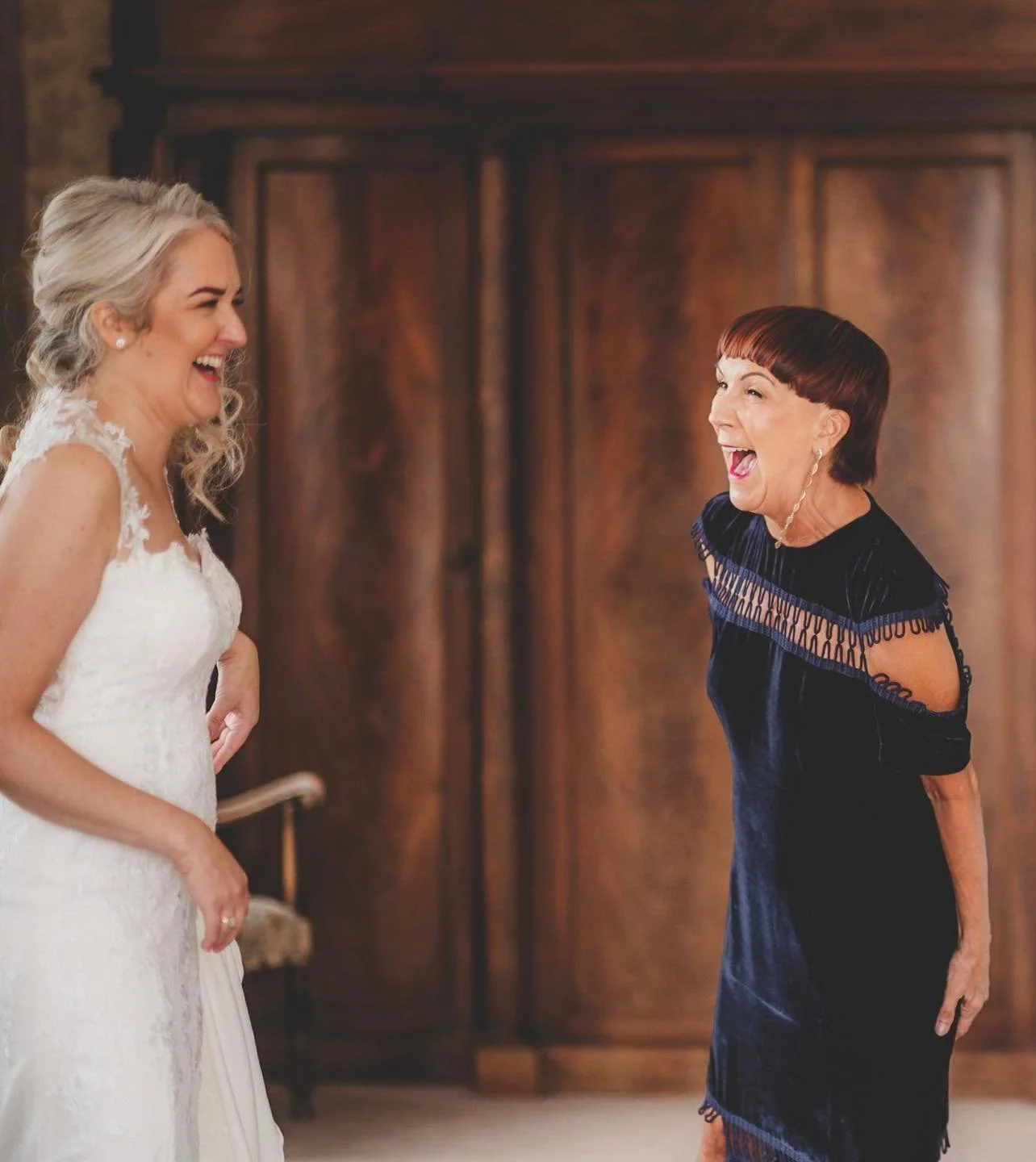 A bride in a white dress laughs joyfully with an older woman in a dark dress. Theyre standing in a warmly lit room with wooden furniture in the background, sharing a candid and joyful moment.
