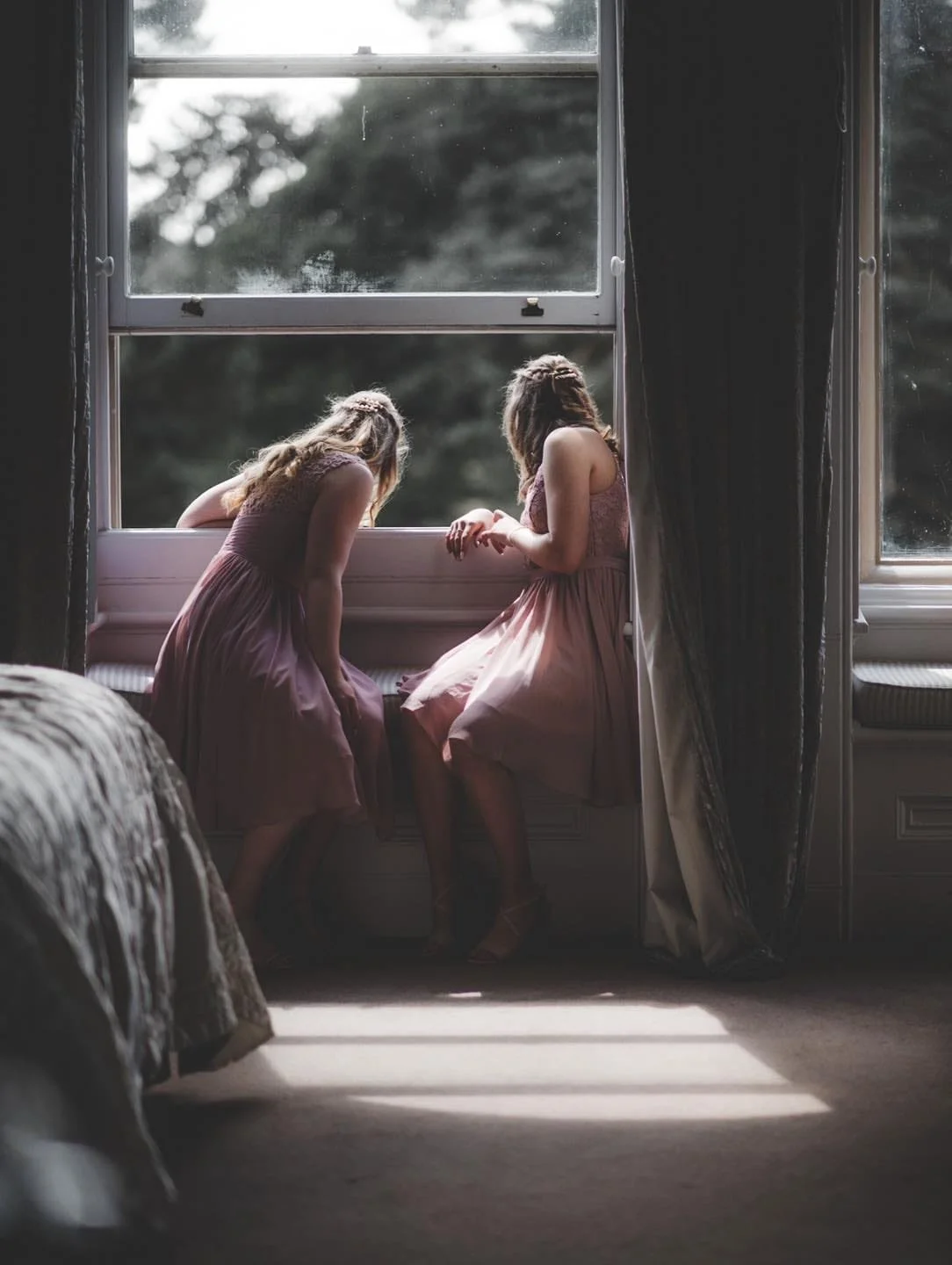 Two young bridesmaids in pastel dresses lean out of a large window, gazing outside. One is seated on the windowsill, while the other bends forward. Soft light from outside bathes the room, creating a serene atmosphere.