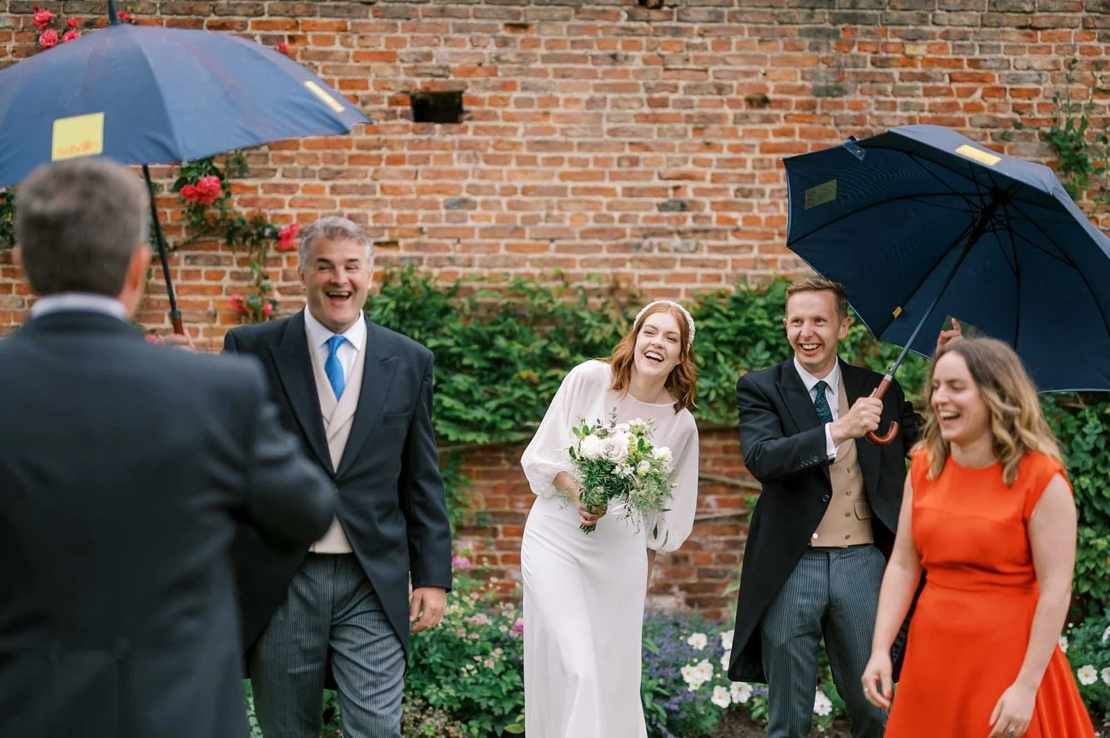 A bride and three other people laugh joyfully in a garden setting, with two holding umbrellas. The background features a brick wall and green foliage. The bride holds a bouquet, and the mood is festive and cheerful.
