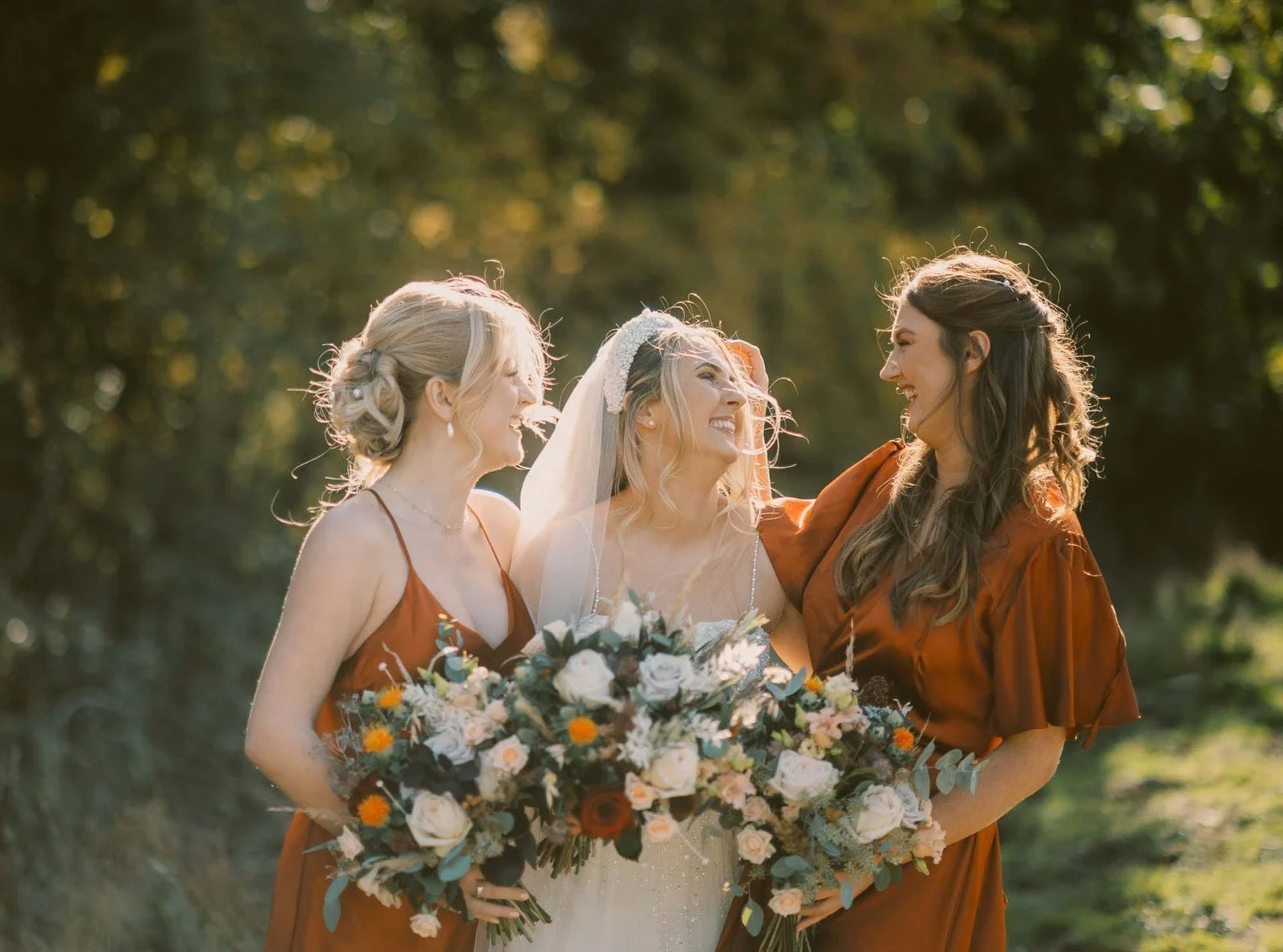 Three smiling women pose outdoors in warm lighting, surrounded by greenery. The bride, in the center, wears a white dress and veil, holding a bouquet. The two bridesmaids wear matching rust-colored dresses, each holding a similar bouquet.