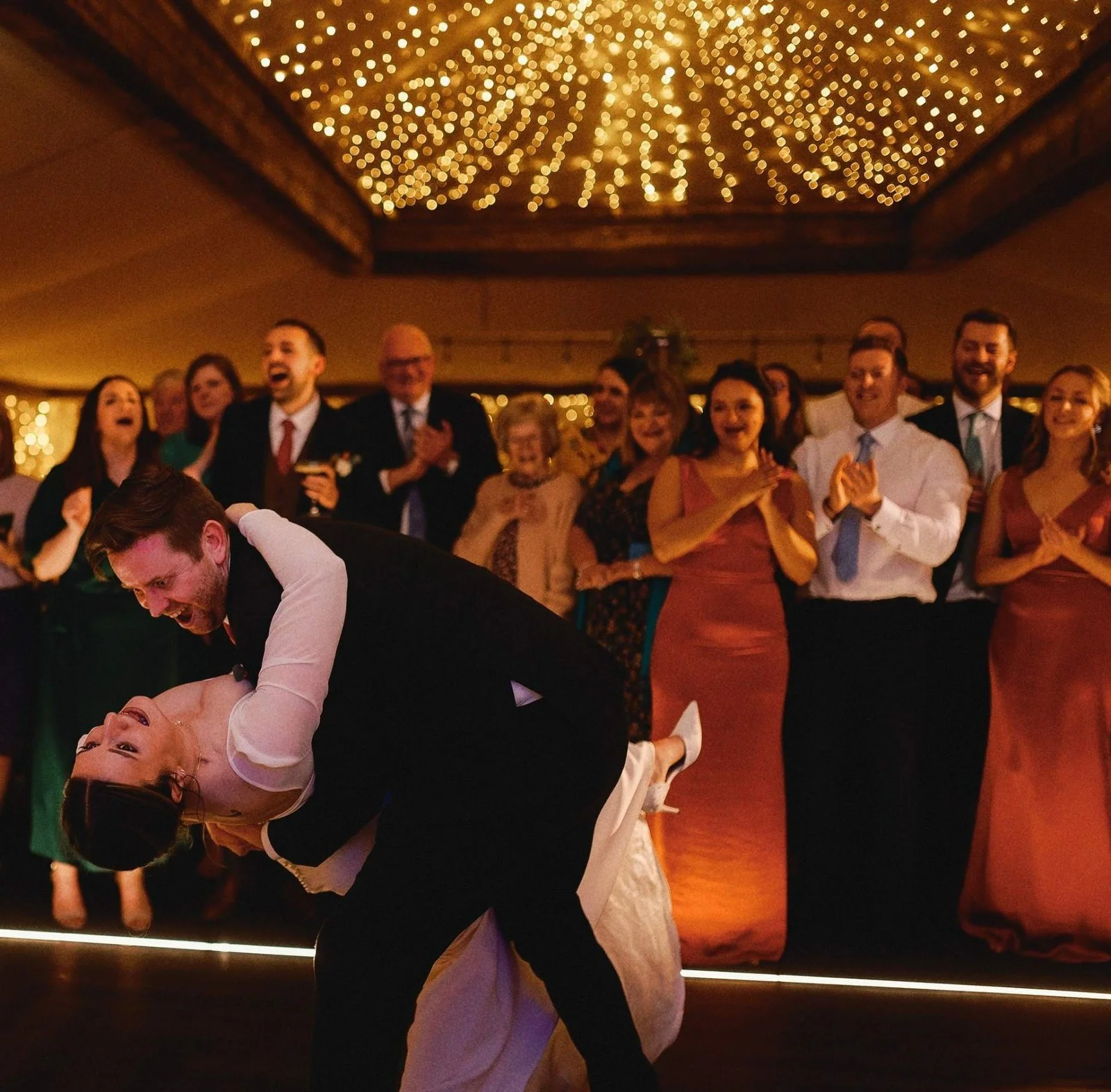 A couple dancing closely at a wedding reception, with a large group of guests clapping and watching happily in the background under a decorated ceiling with string lights.