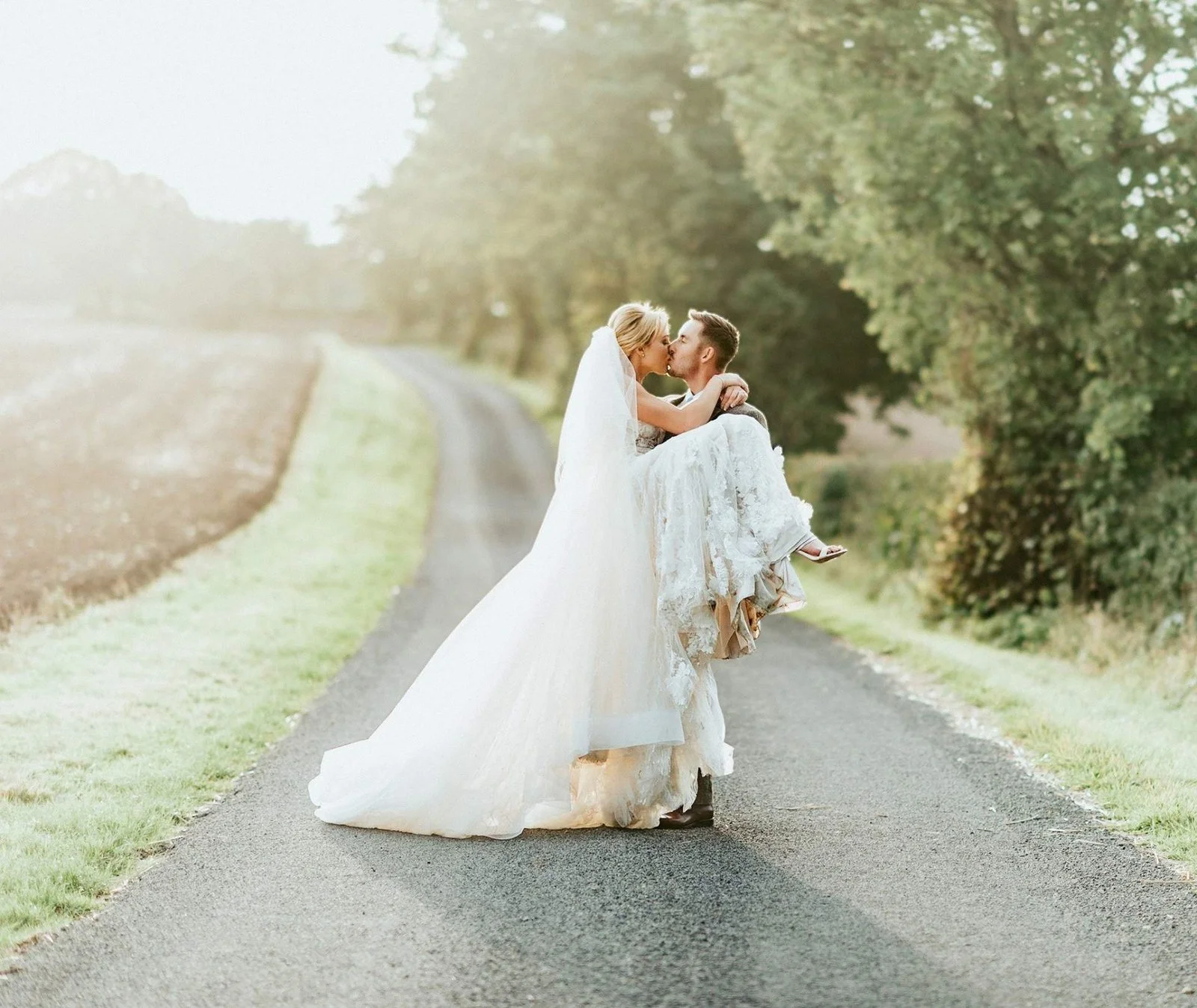 A couple in wedding attire kissing on a rural road surrounded by green trees.