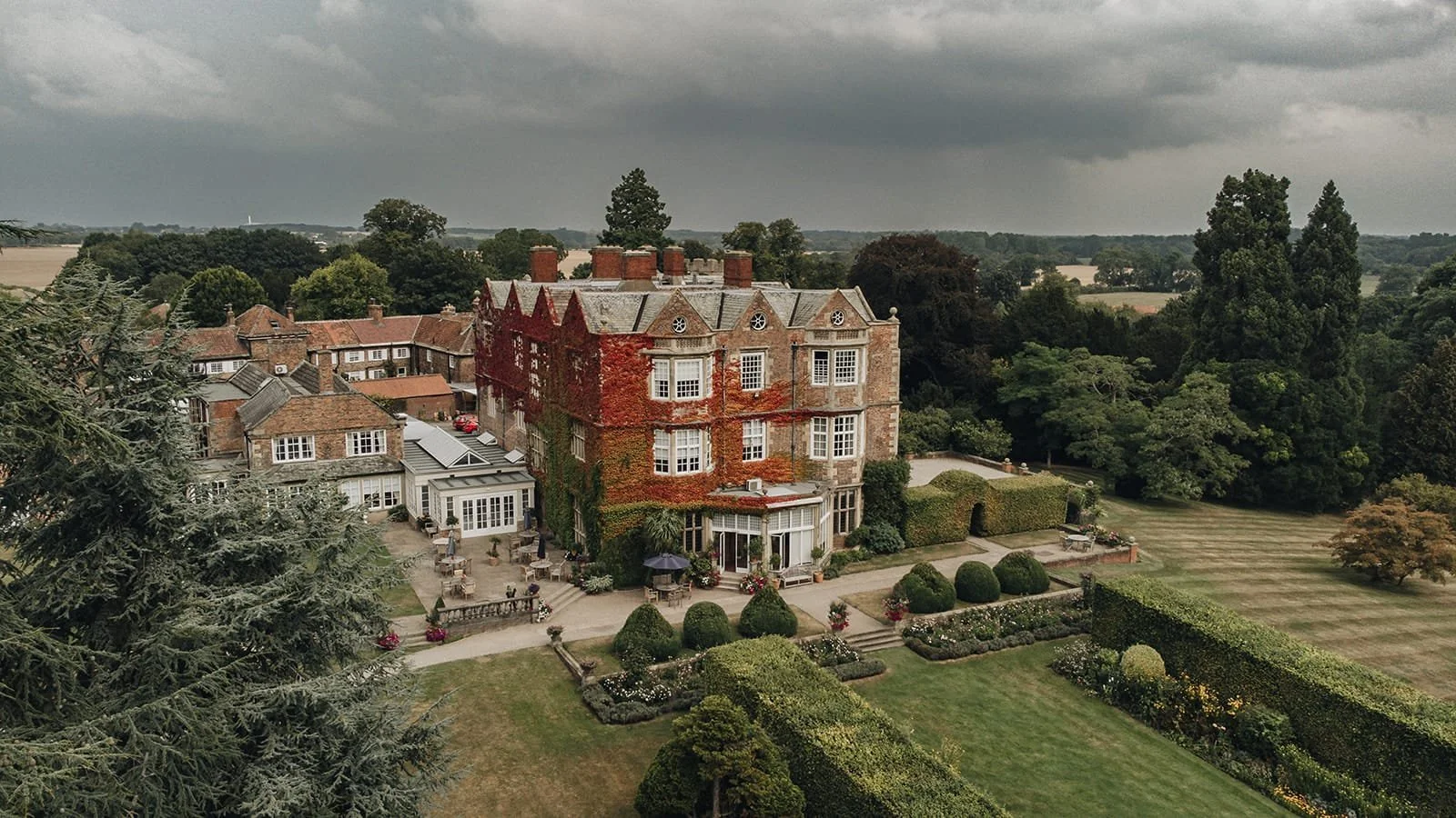 A large historic mansion with brick and ivy-covered walls, surrounded by a well-maintained garden and hedges, under a cloudy sky.