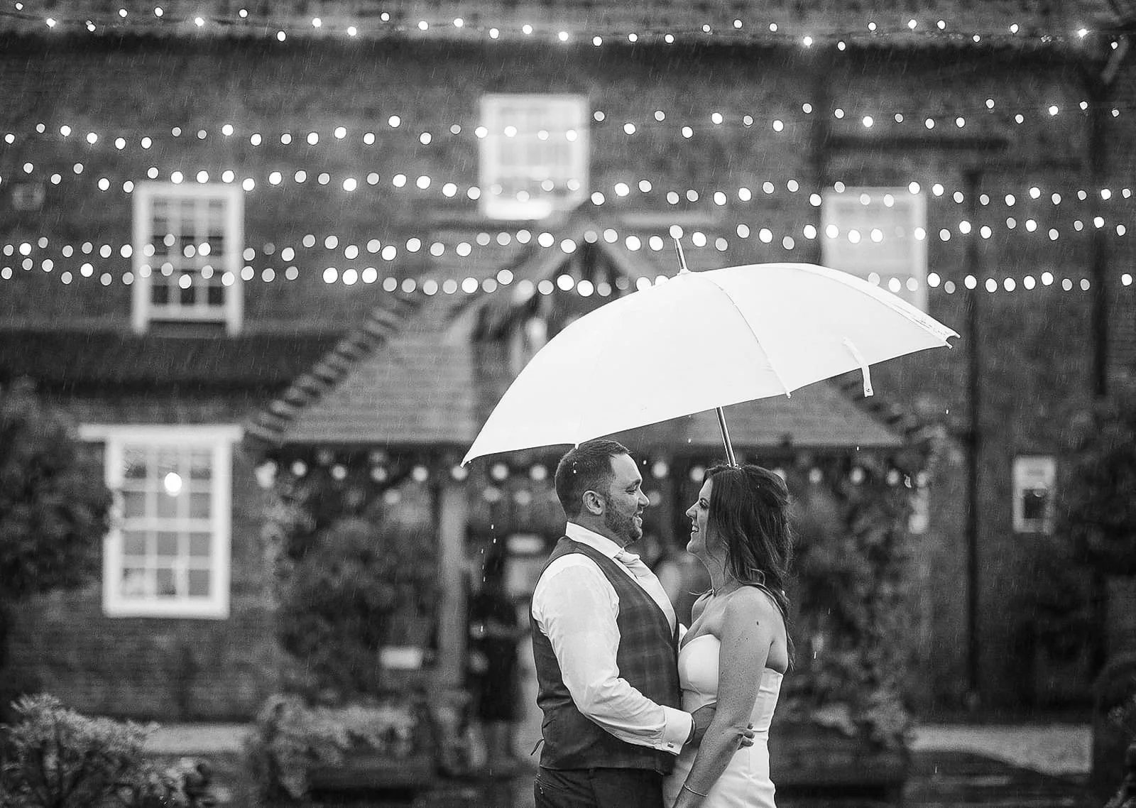 A couple in wedding attire is standing close together under an umbrella, sharing a joyful moment in the rain at night. String lights are hanging above them in front of a rustic brick building with windows and greenery.