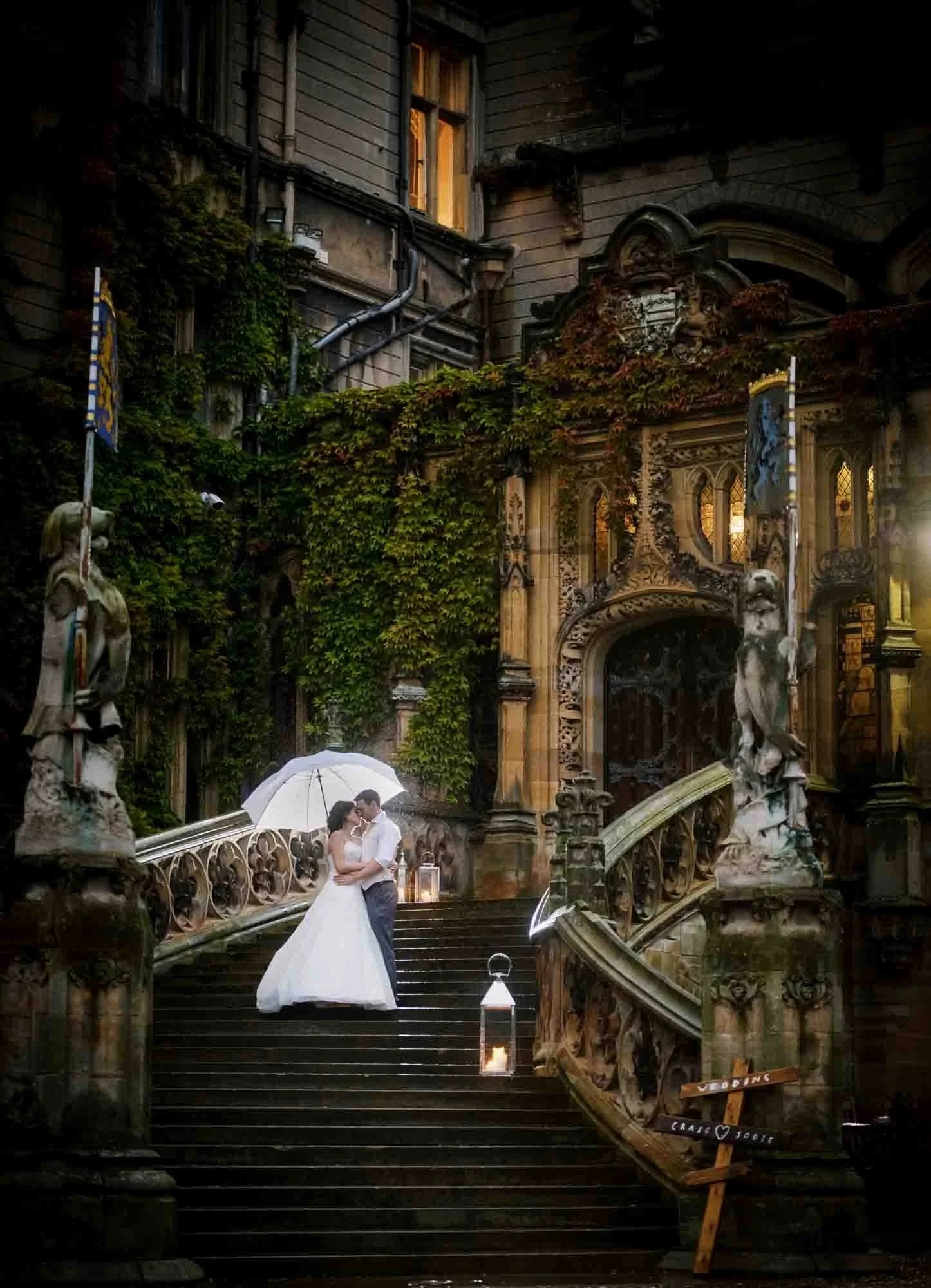 A bride and groom standing close together, sharing a kiss under a white umbrella on a grand, ornate staircase with candles and lanterns around them, surrounded by ivy-covered walls and historic architecture.