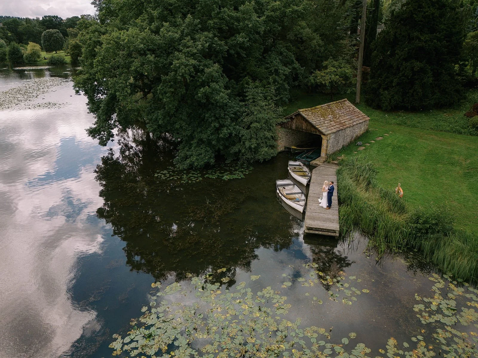 A bride and groom standing on a dock next to a pond, with three small boats beside them and a boathouse nearby. The scene is surrounded by lush greenery and trees.
