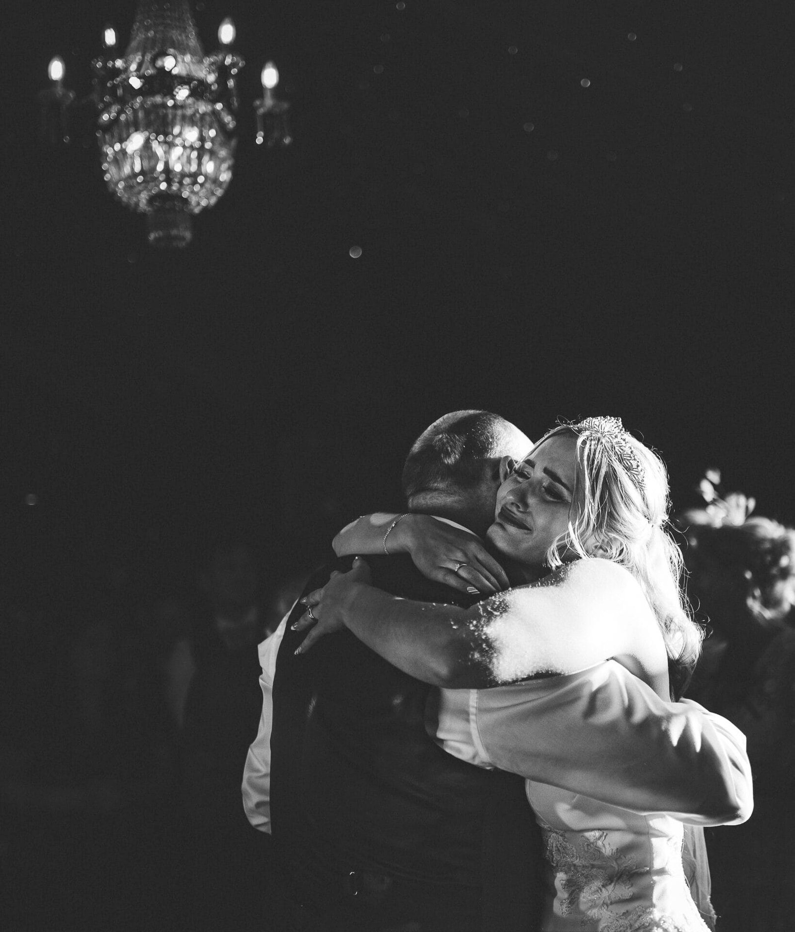 Black and white photo of a couple embracing, likely at a wedding reception, with a chandelier hanging from the ceiling in the background, inside in the grounds of Norwood Park