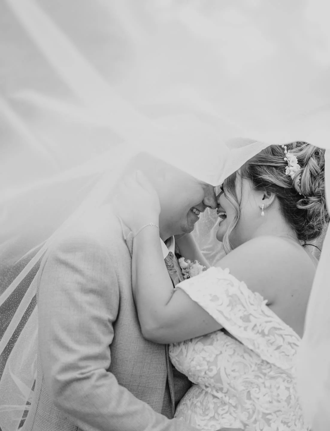 Black and white wedding photo of a bride and groom with their foreheads touching, under a veil, smiling and holding each other, in the grounds of Norwood Park
