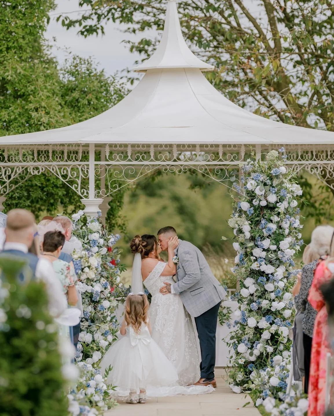 A bride and groom kissing at an outdoor wedding ceremony under a white gazebo decorated with flowers, with guests watching, taken by a Norwood Park wedding photographer