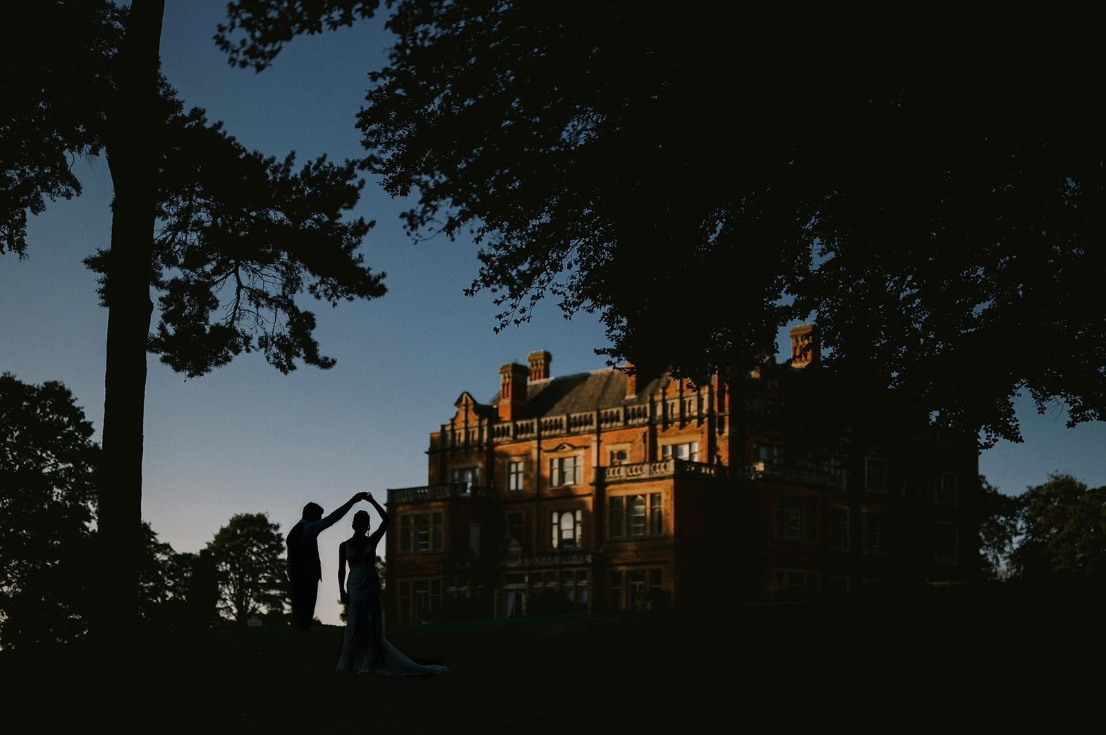 Silhouette of a couple dancing outside in front of a large, historic mansion with a sunset or dusk sky. Rossinghall wedding photographer taken as the sun sets on a wedding day