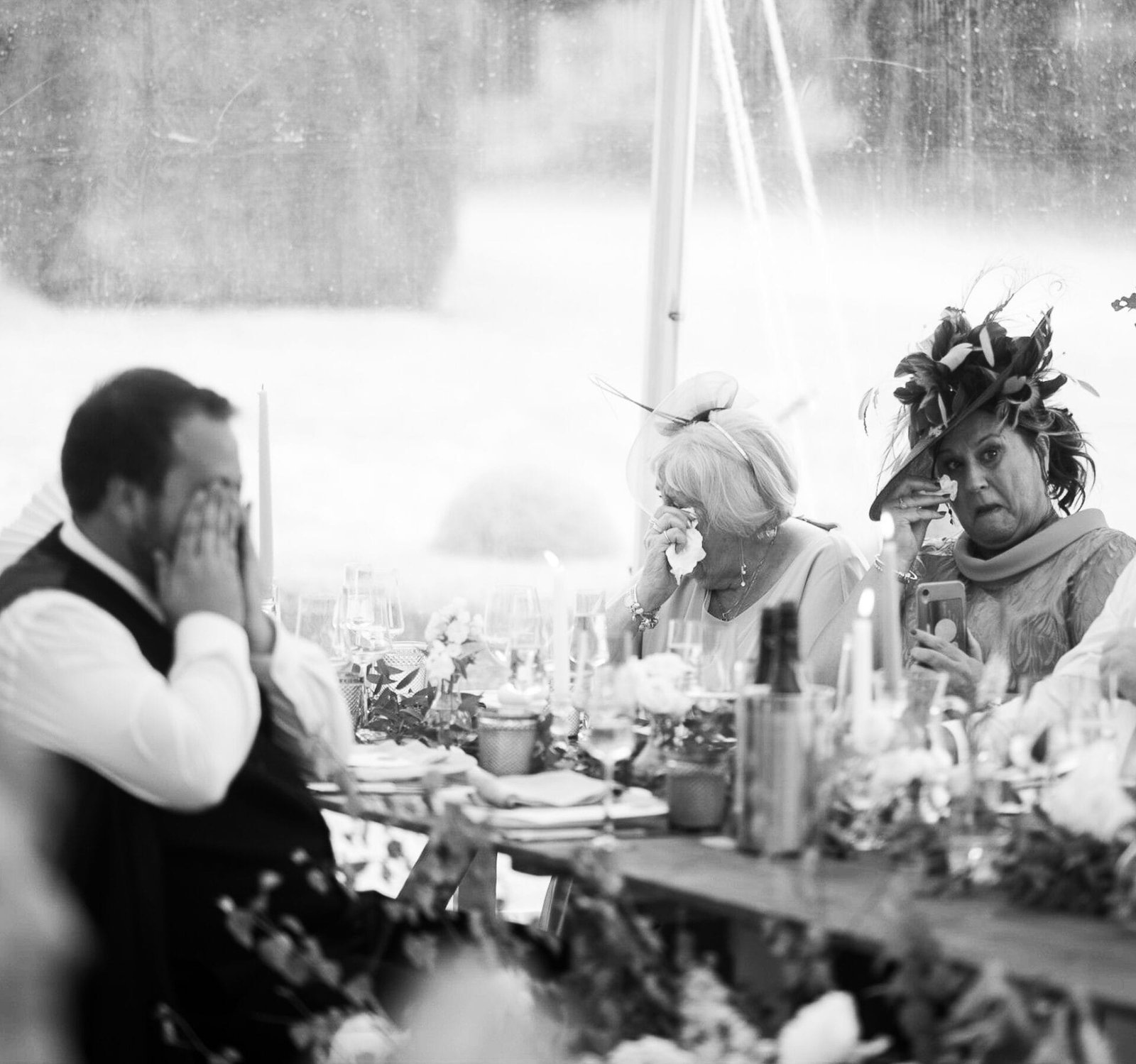 A black and white photo of three people sitting at a table. The person on the left has their face covered with their hands. The middle woman, wearing a hat and glasses, is wiping tears from her eyes. The woman on the right, also wearing a hat, is hol