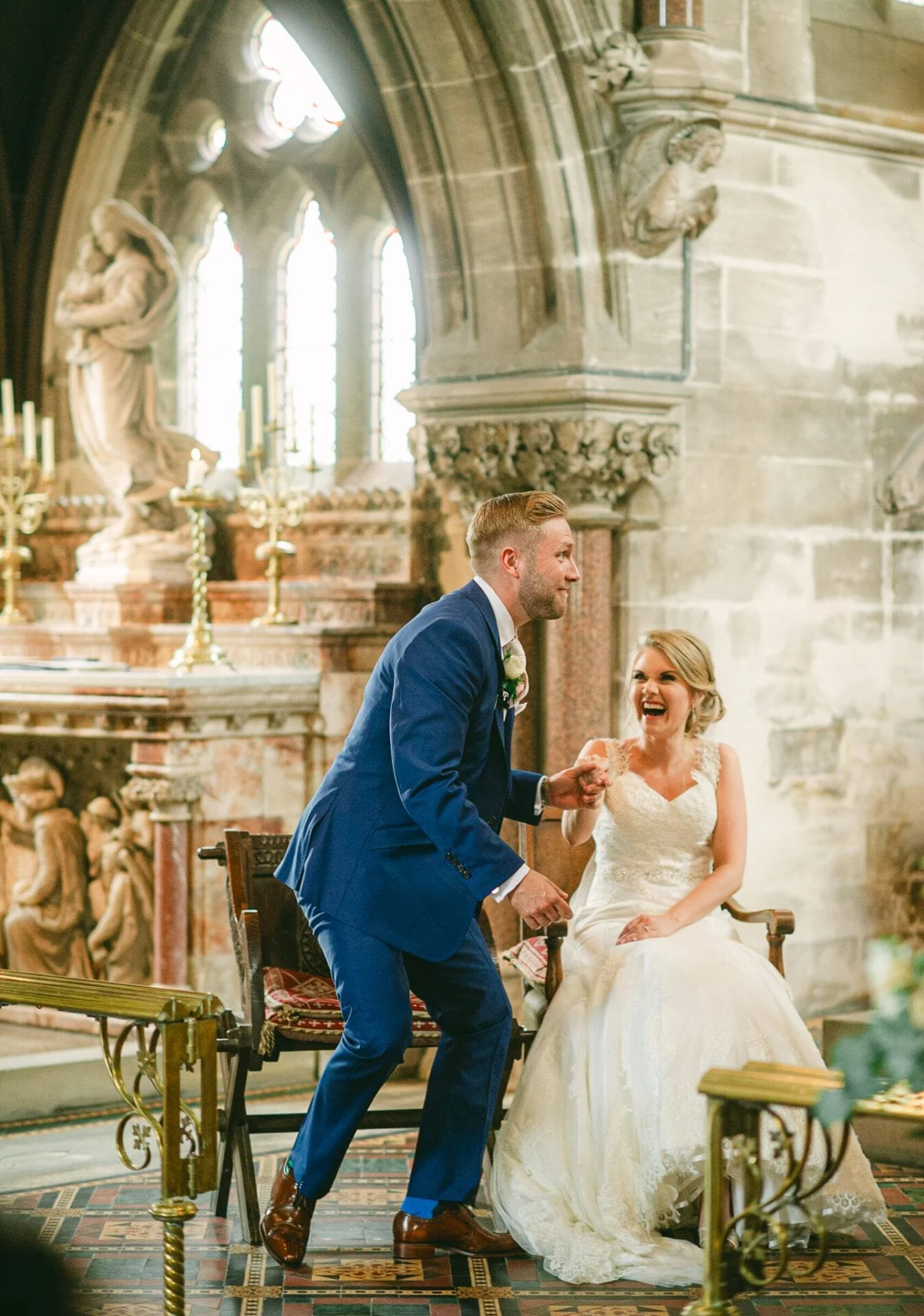 A bride and groom celebrating during a wedding ceremony inside a historic church with stained glass windows and stone sculptures, with the groom holding the bride's hands and both laughing.