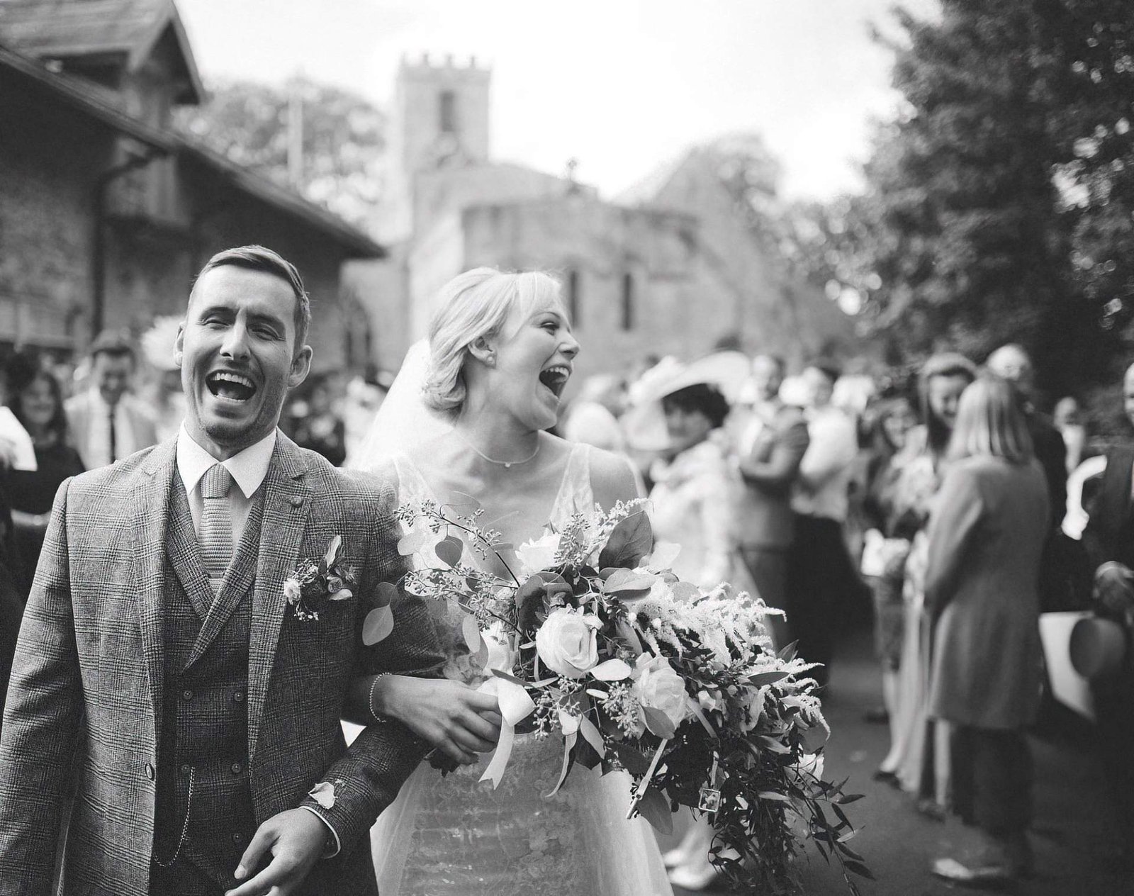 Black and white photo of a newlywed couple laughing and walking outdoors, surrounded by wedding guests. The groom wears a plaid suit with a boutonnière, and the bride holds a large bouquet with foliage, wearing a lace dress and veil. The background s
