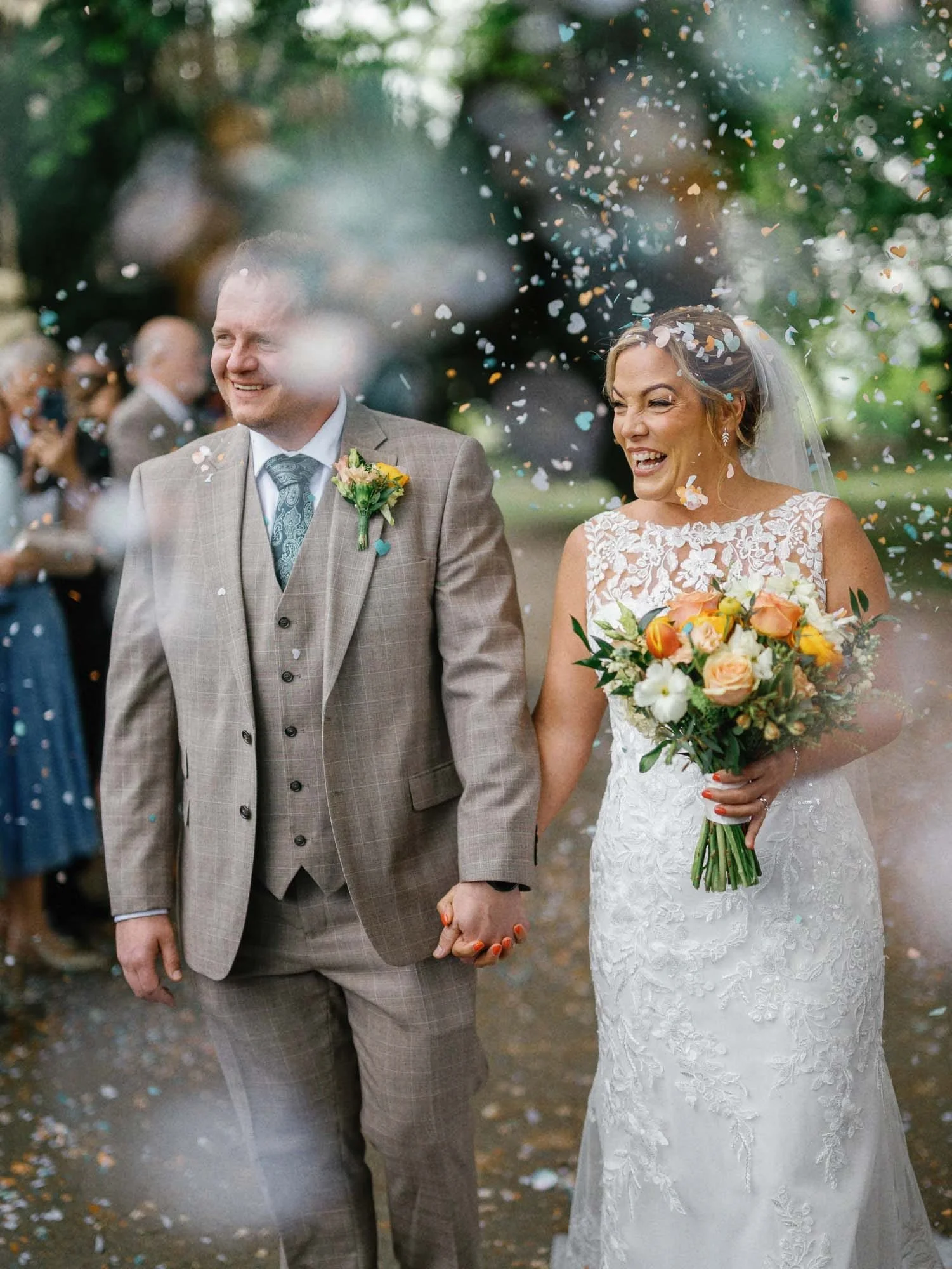 Bride and groom holding hands at their wedding, surrounded by guests and falling confetti, outdoors in a garden setting.