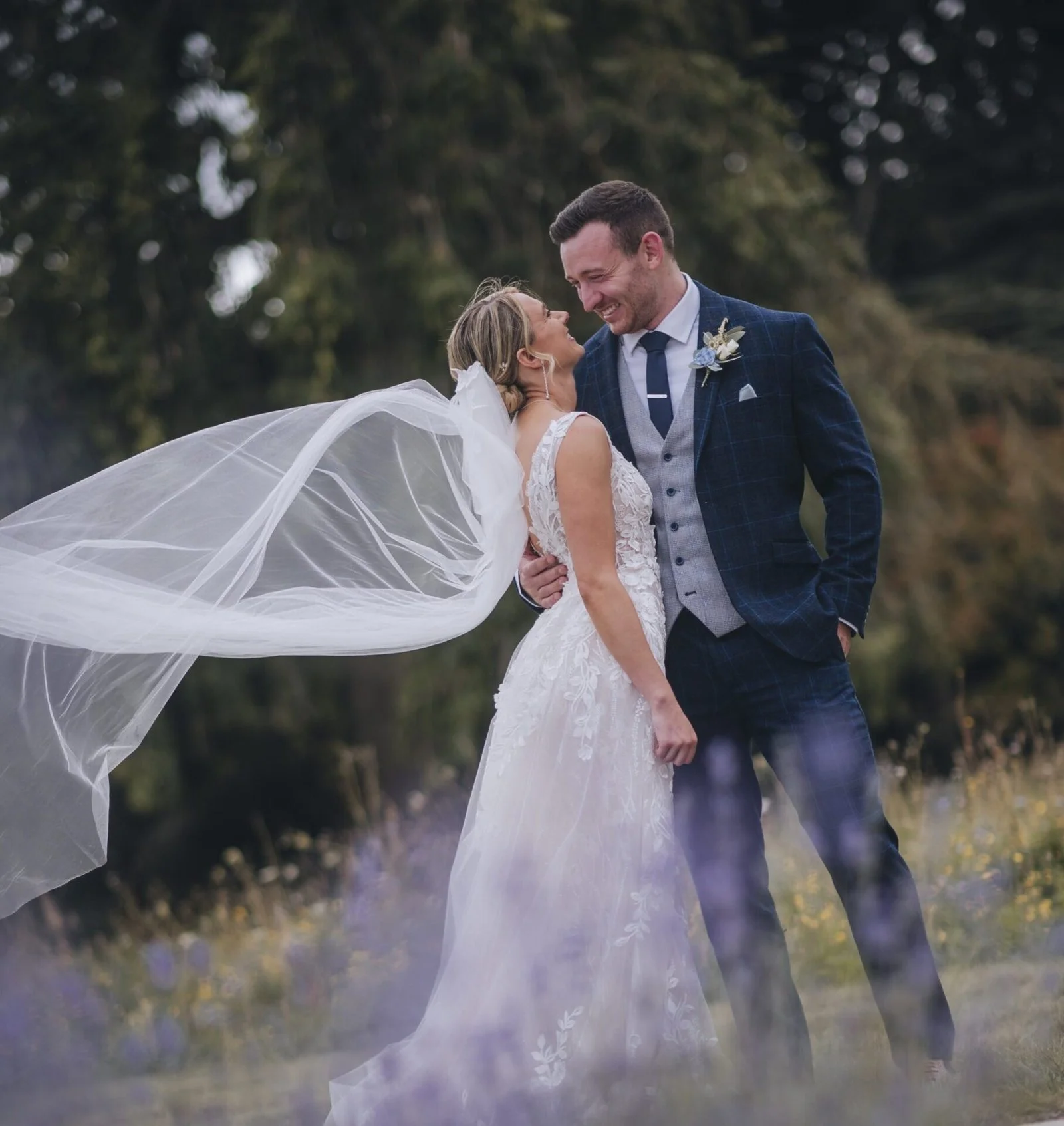 Couple in wedding attire smiling and looking at each other outdoors with trees in the background, in the grounds of Norwood Park