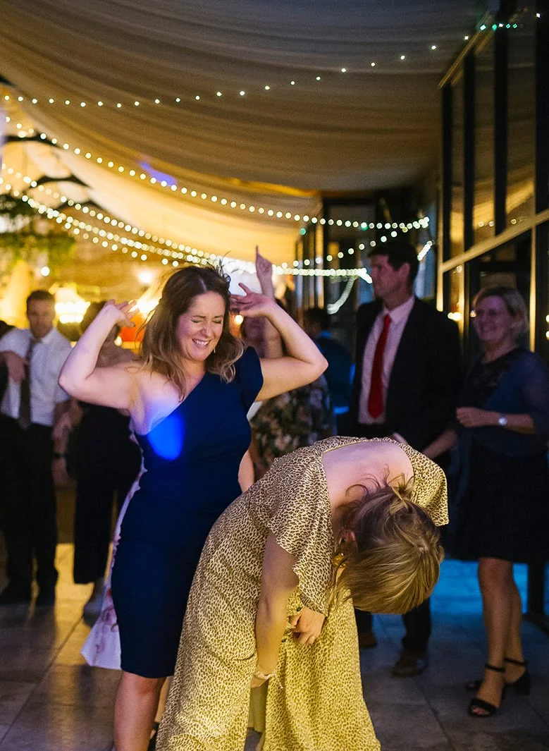 People dancing and enjoying themselves at a celebration or party under string lights, with two women in foreground, one in a blue dress dancing with a big smile and the other in a yellow animal print dress bent over. Yorkshire wedding photo
