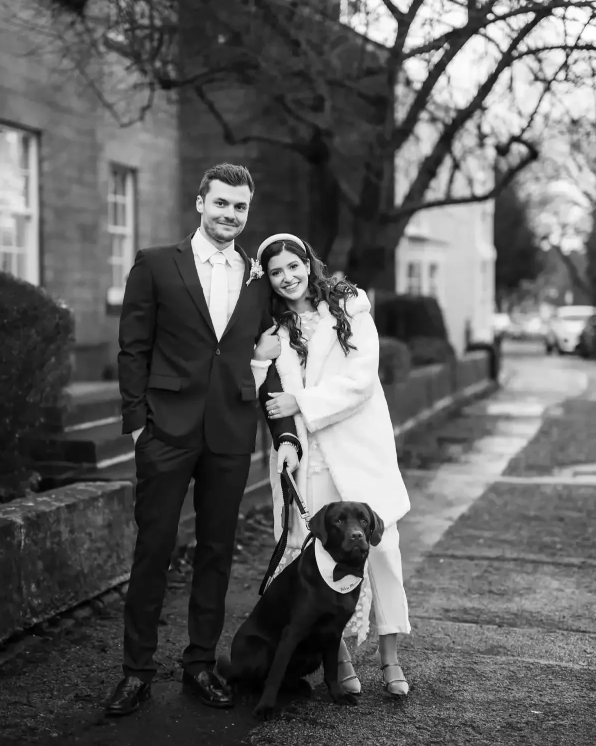 Black and white portrait of a happy couple and their black Labrador Retriever wearing a bowtie outside harrogte registrar office on a wedding day