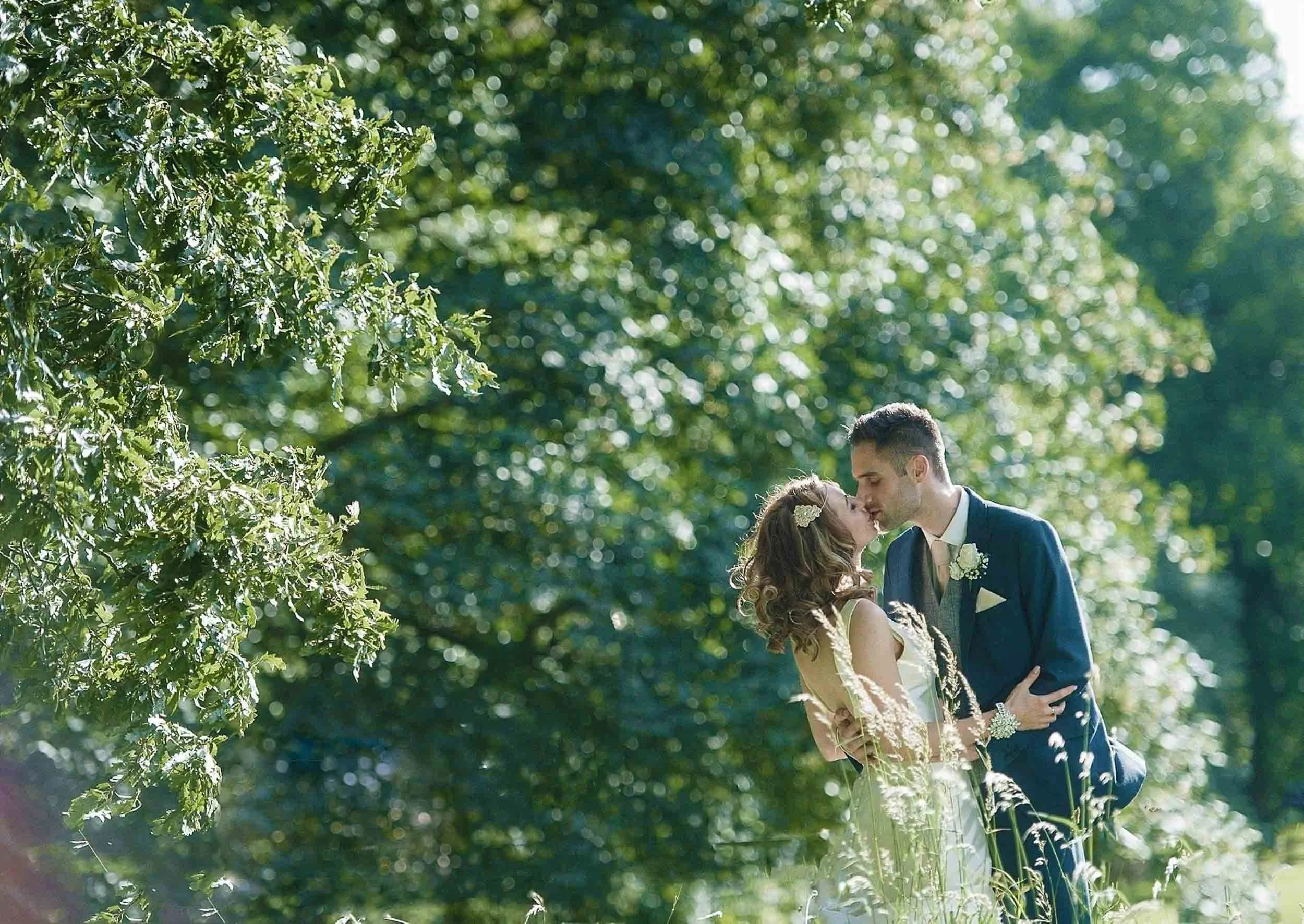 A bride and groom sharing a wedding kiss outdoors, surrounded by green trees and sunlight.