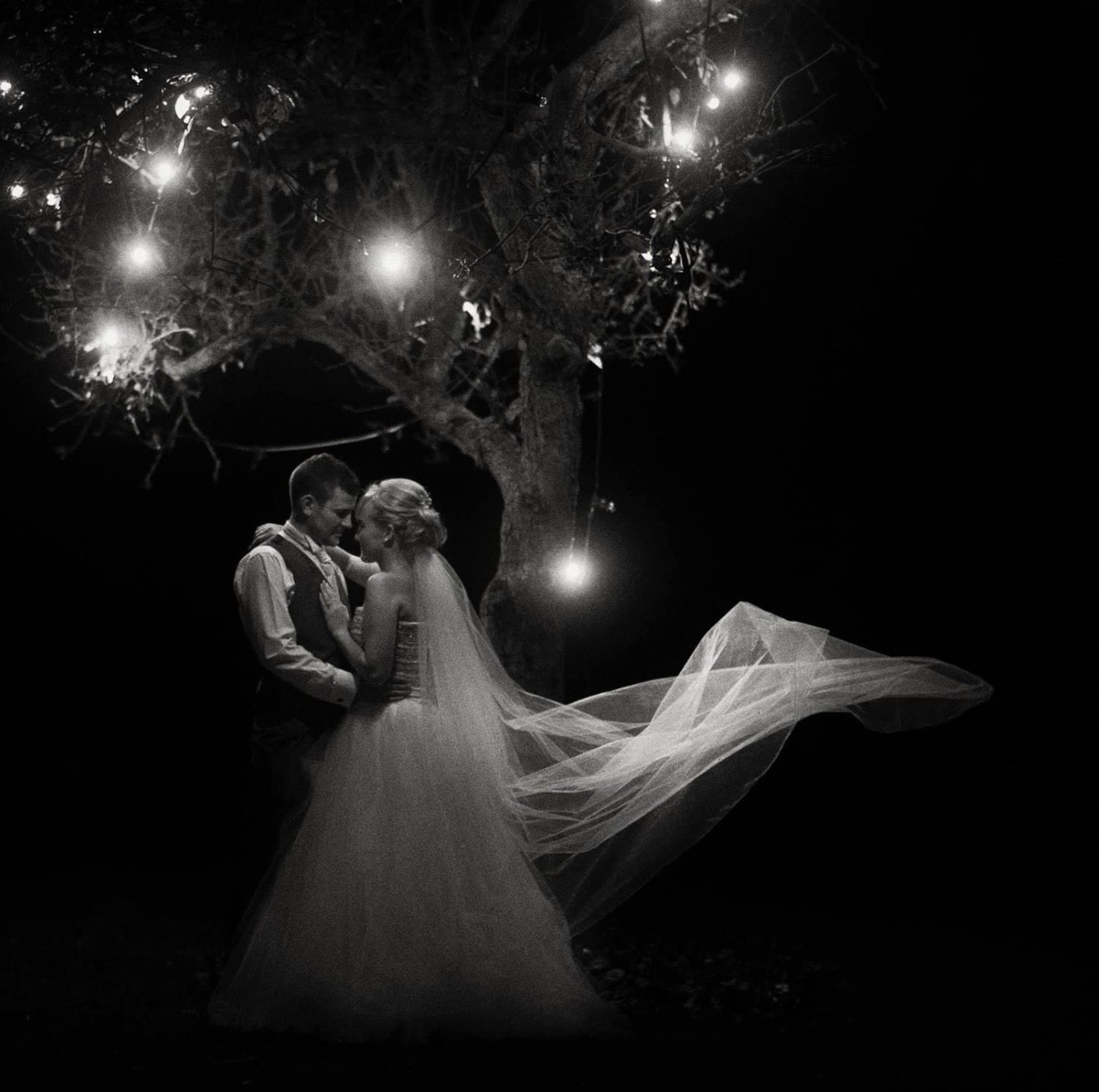 A black-and-white photo of a bride and groom embracing under a lit tree at night.