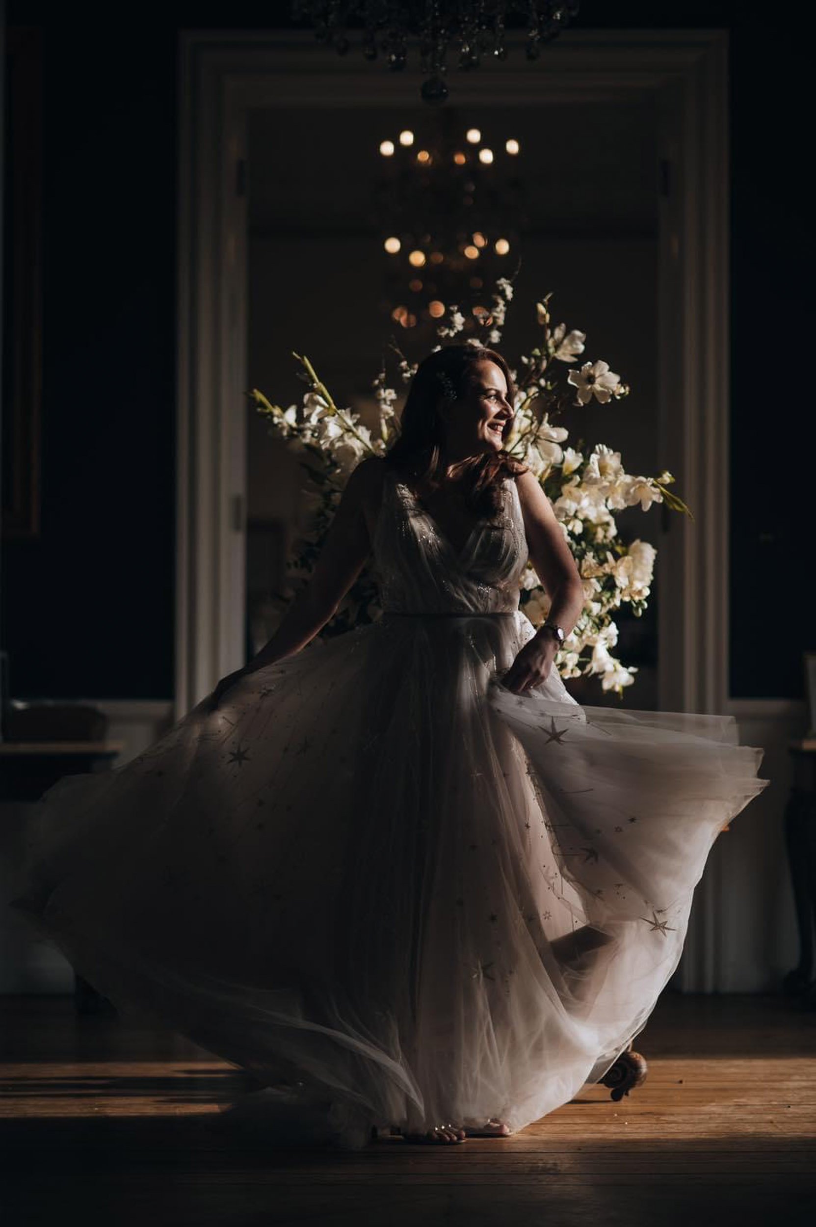 A woman in a flowing, star-embellished gown spinning and smiling on a wooden floor with a floral arrangement and chandelier in the background.