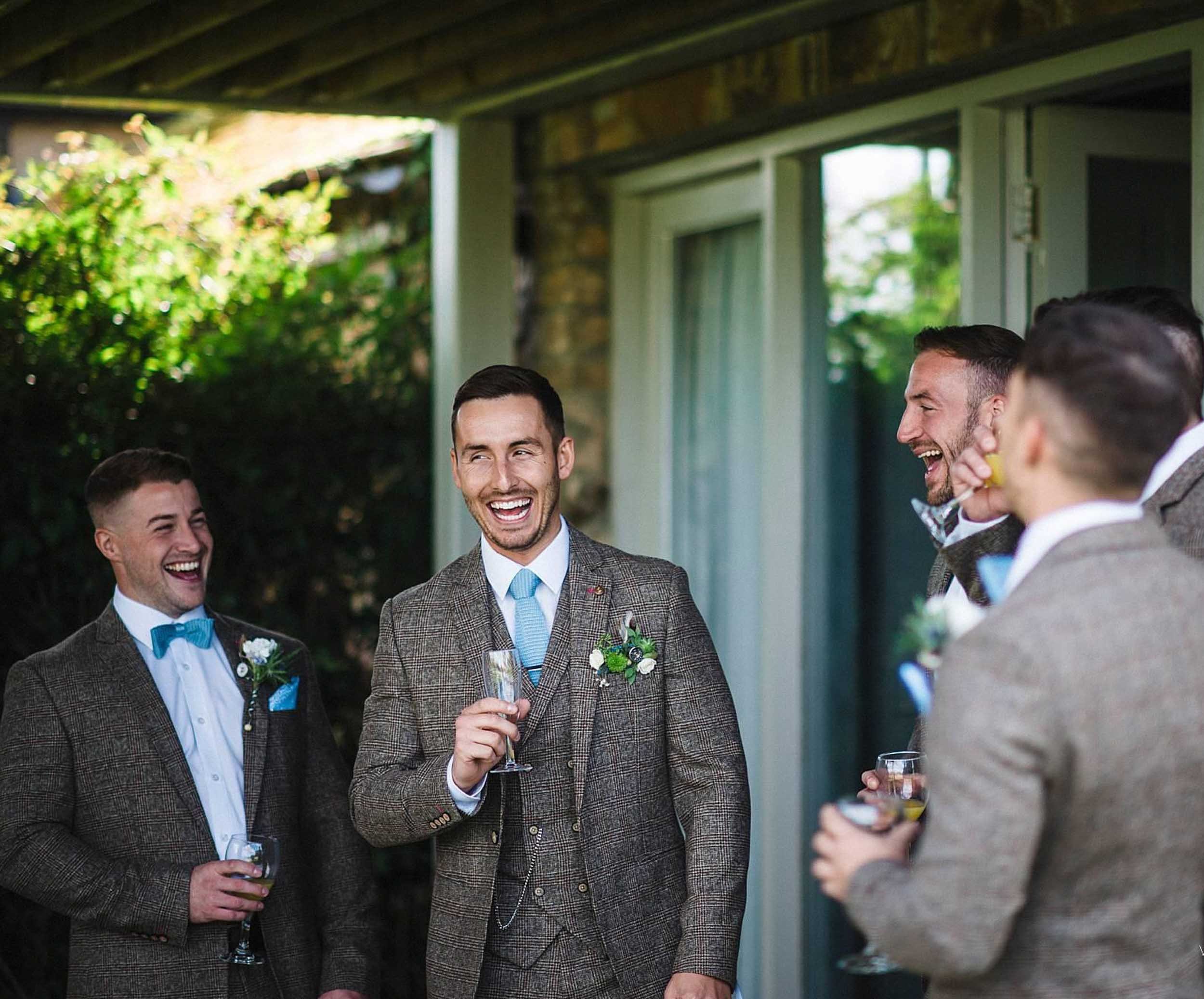 Group of men dressed in suits and bow ties, smiling and laughing while holding drinks outdoors near a house with trees in the background.