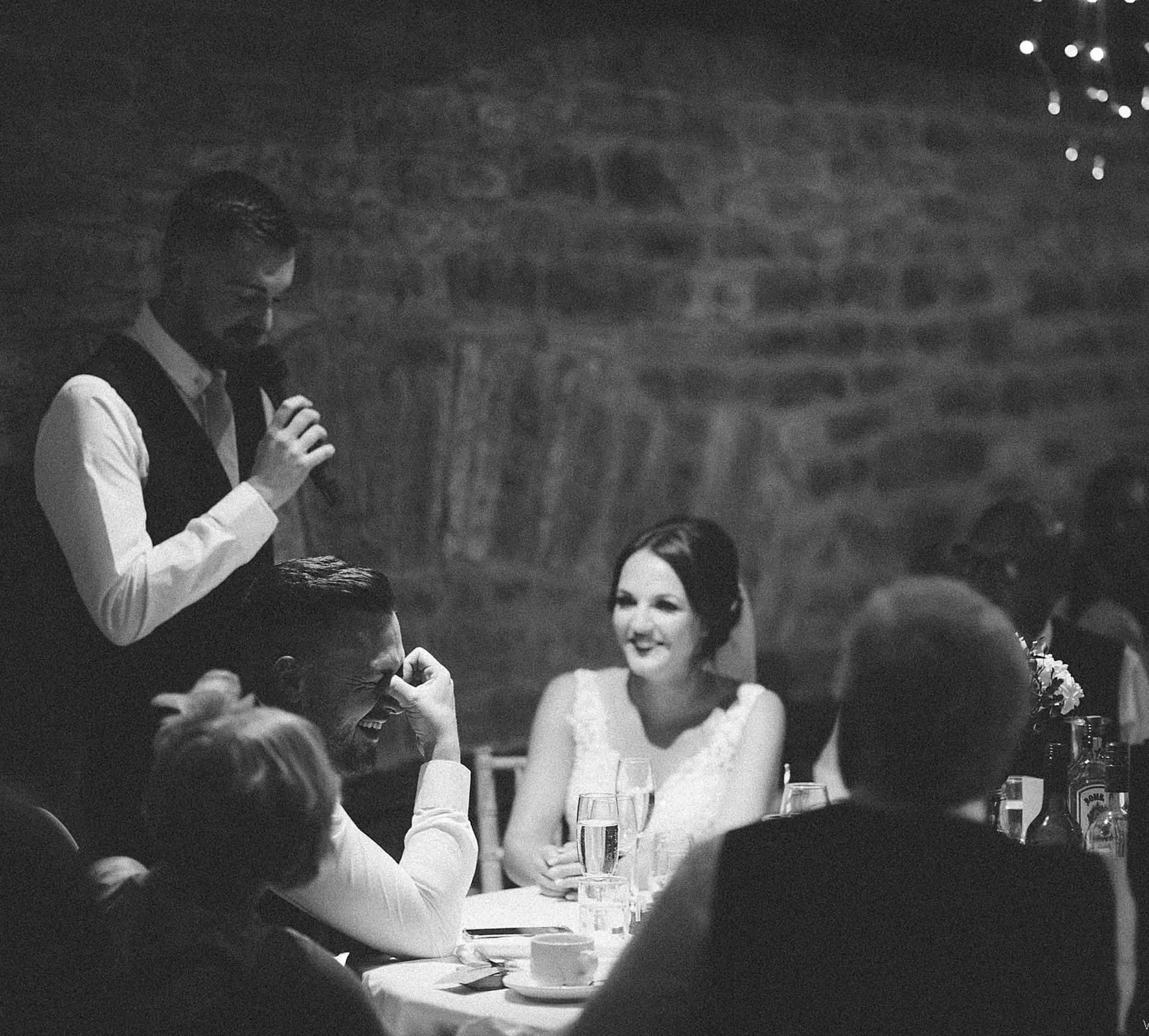 A group of people at a wedding reception, with a woman in a wedding dress sitting at a table, smiling, and a man sitting next to her laughing. A man standing and speaking into a microphone is also present, with a brick wall in the background and glas