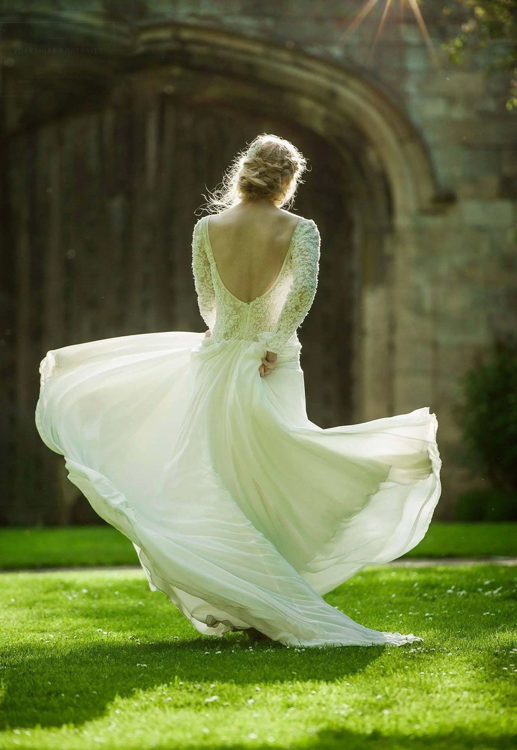 A woman in a flowing wedding gown spinning in a grassy field with a stone archway in the background, back facing the camera, sunlight shining.