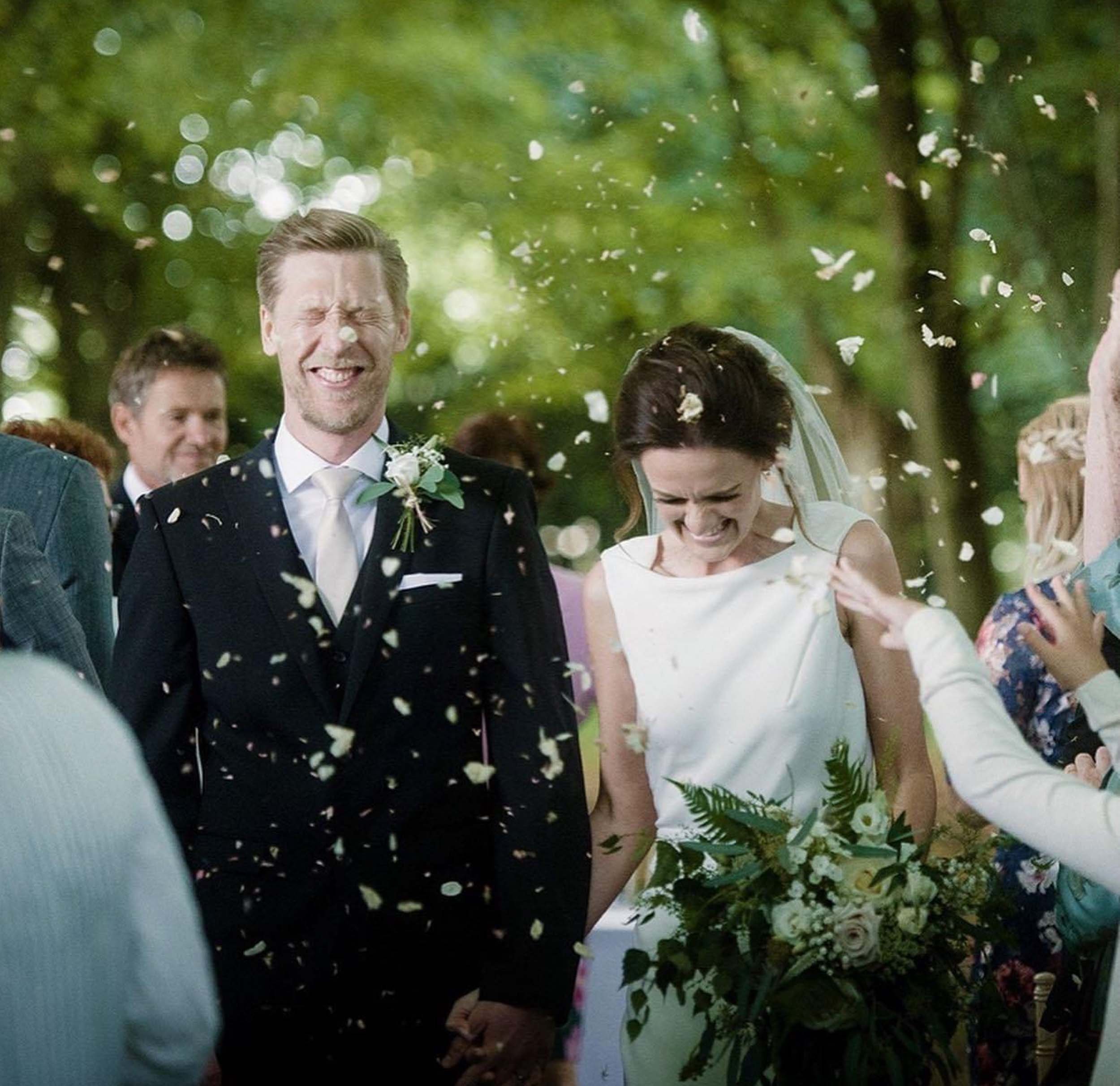 A bride and groom celebrating their wedding outdoors, smiling and laughing as family and friends throw rice or flower petals in the air.