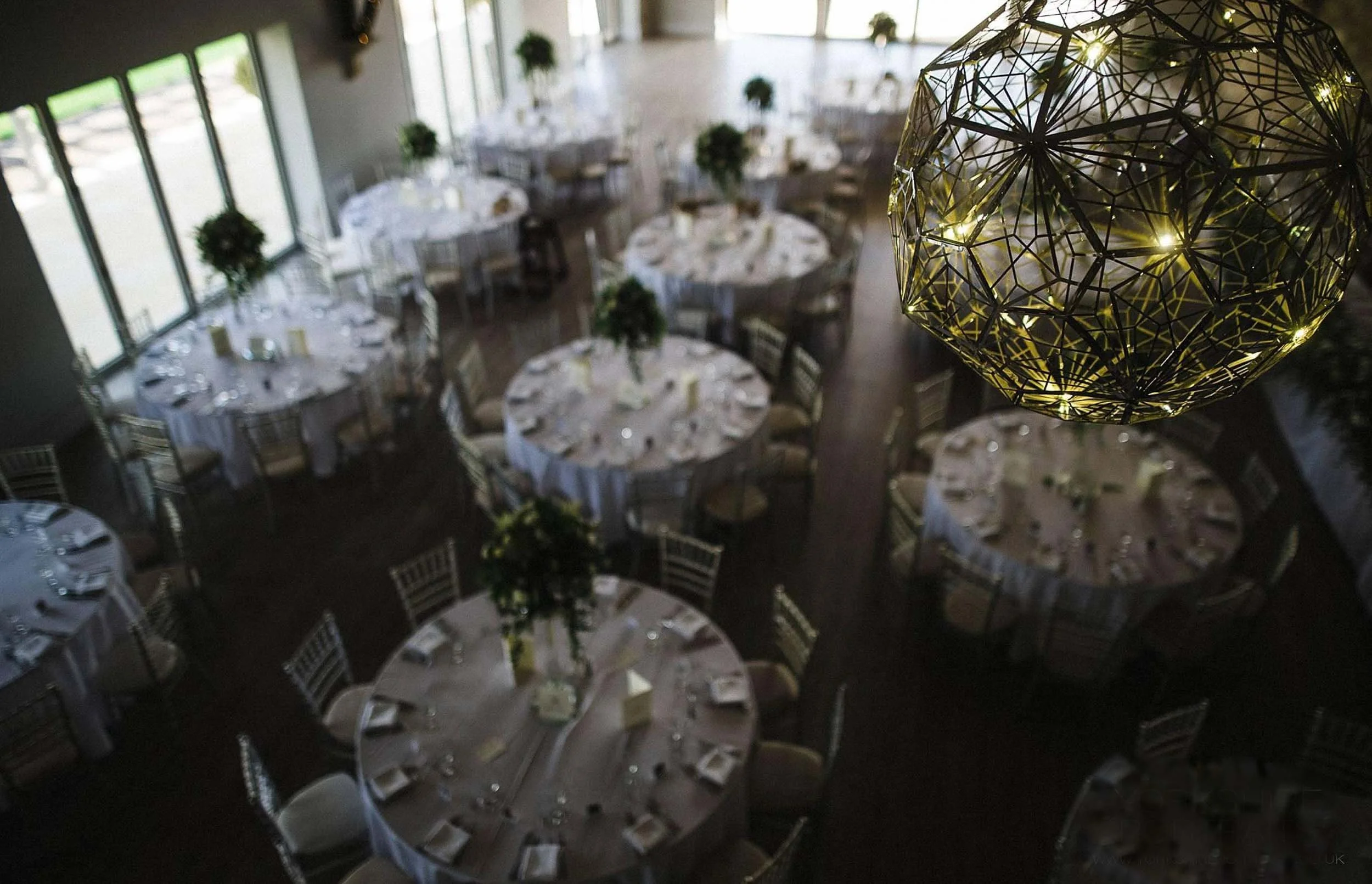 Elegant event hall with round tables decorated with white tablecloths and floral centerpieces, large windows letting in natural light, and a large decorative chandelier with golden geometric pattern overhead.