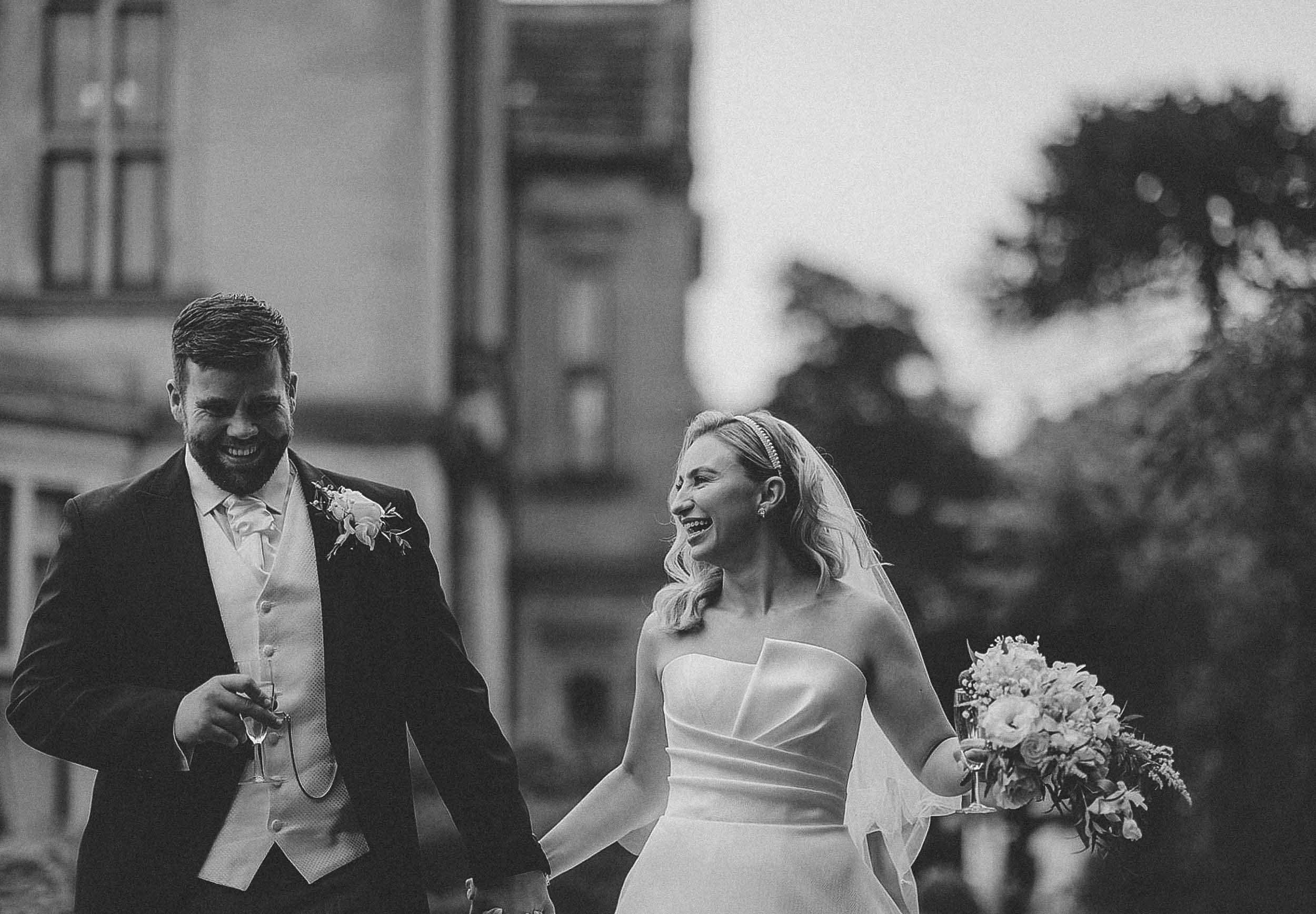 Black and white photo of a happy bride and groom holding hands outdoors, both smiling and laughing. The bride holds a bouquet of flowers and the groom holds a glass of champagne.