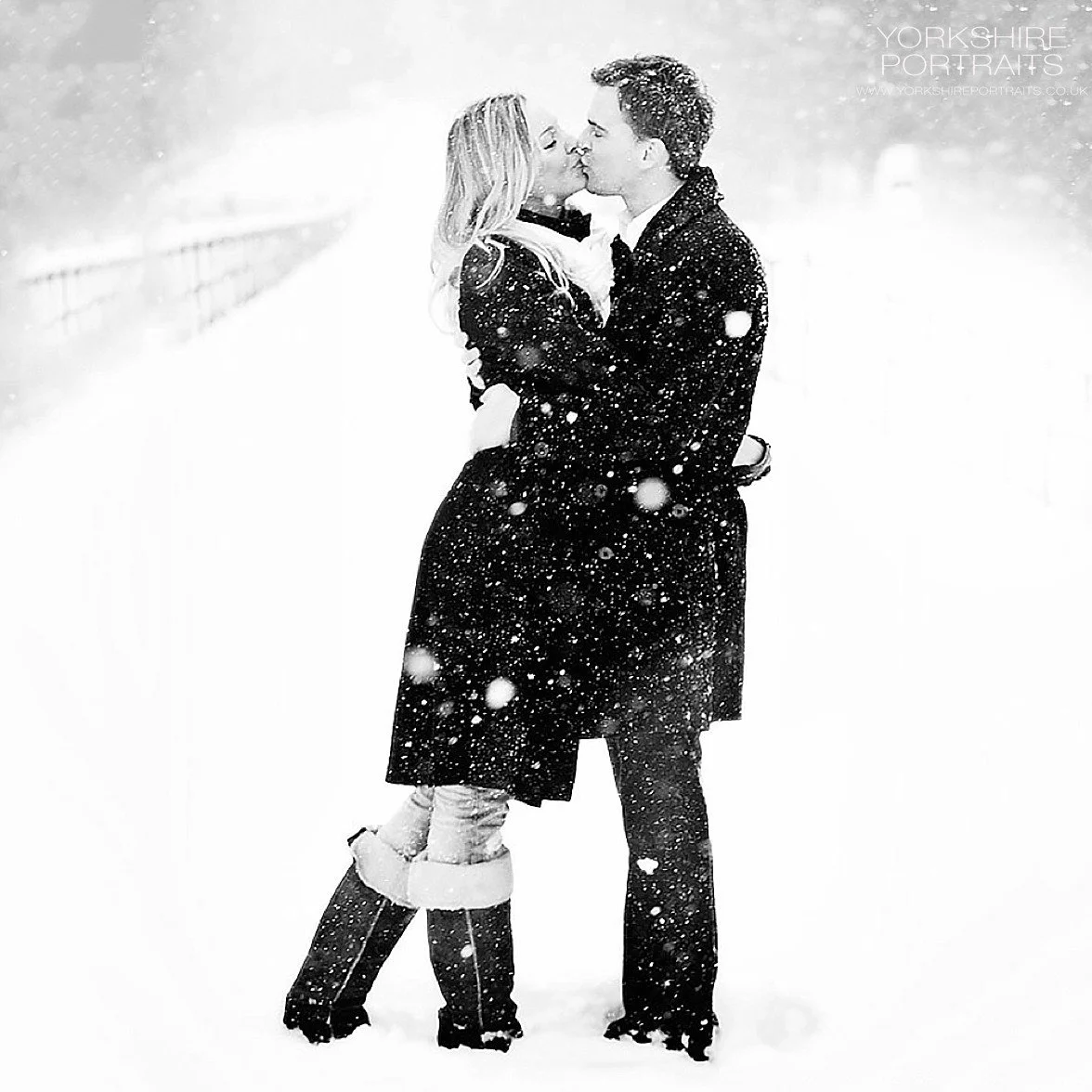 A couple kissing in the snow with snow falling around them, dressed in dark winter clothing, outdoors in a snowy landscape.