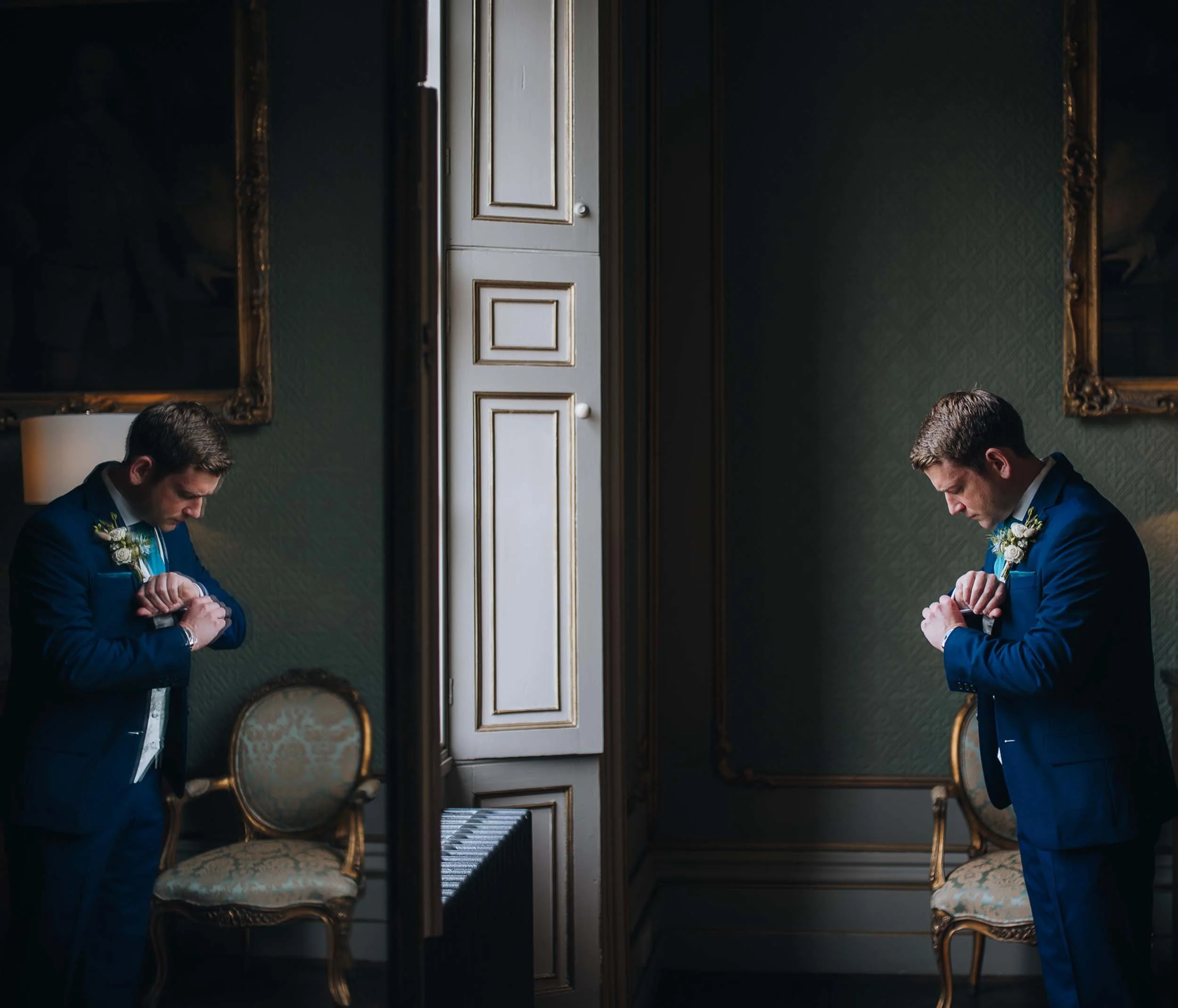 A man in a navy suit checking his watch while looking down in a room with green wallpaper, gold framed mirrors, and a chair, reflected in a room mirror.