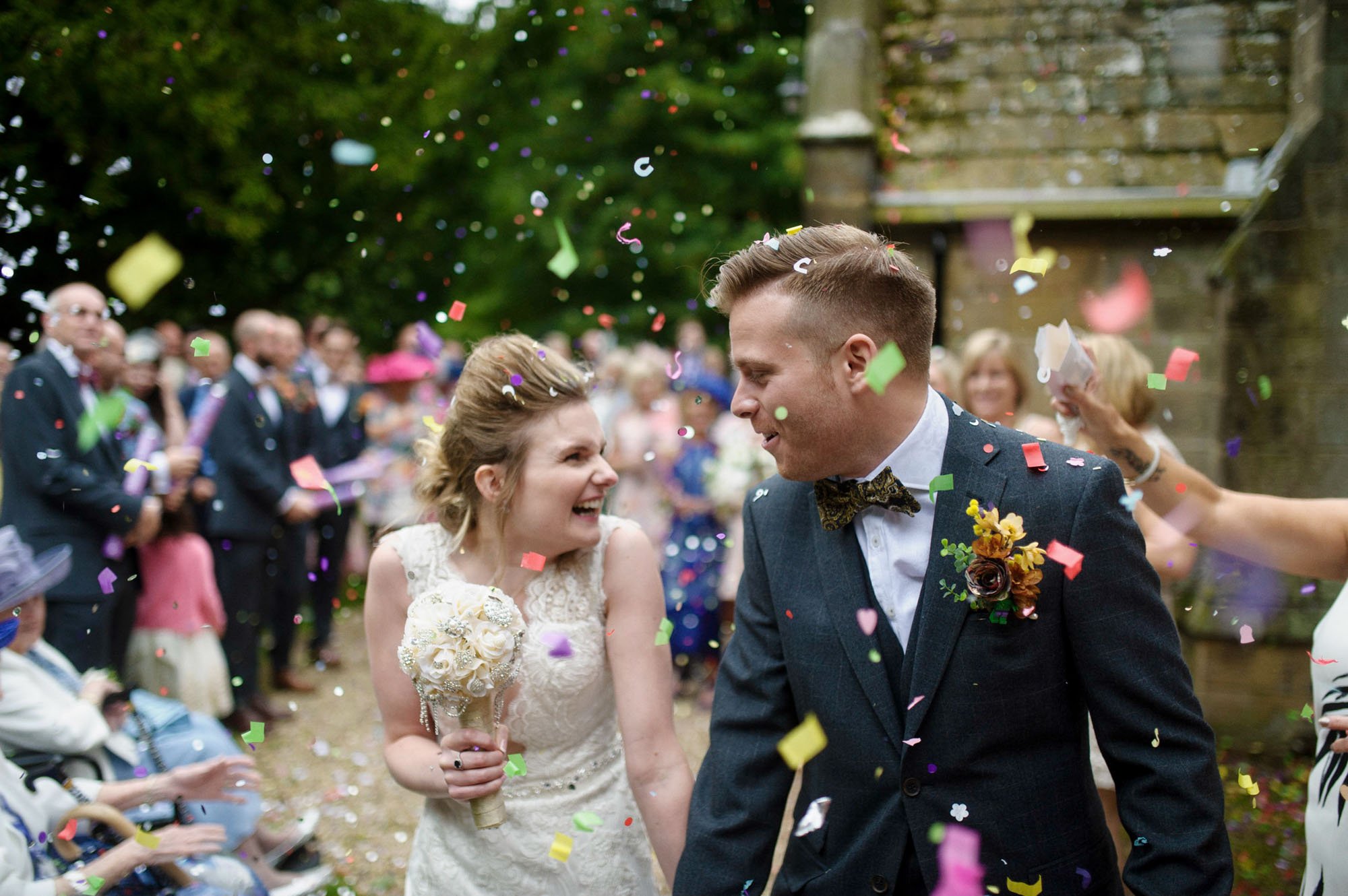 A bride and groom happily walking through falling confetti at wedding with guests in the background.
