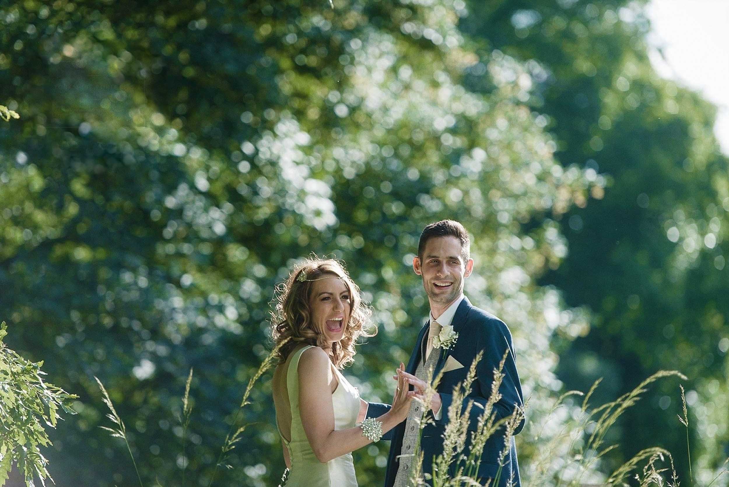 A happy bride and groom standing outdoors in a natural setting with trees and greenery, smiling and looking at the camera