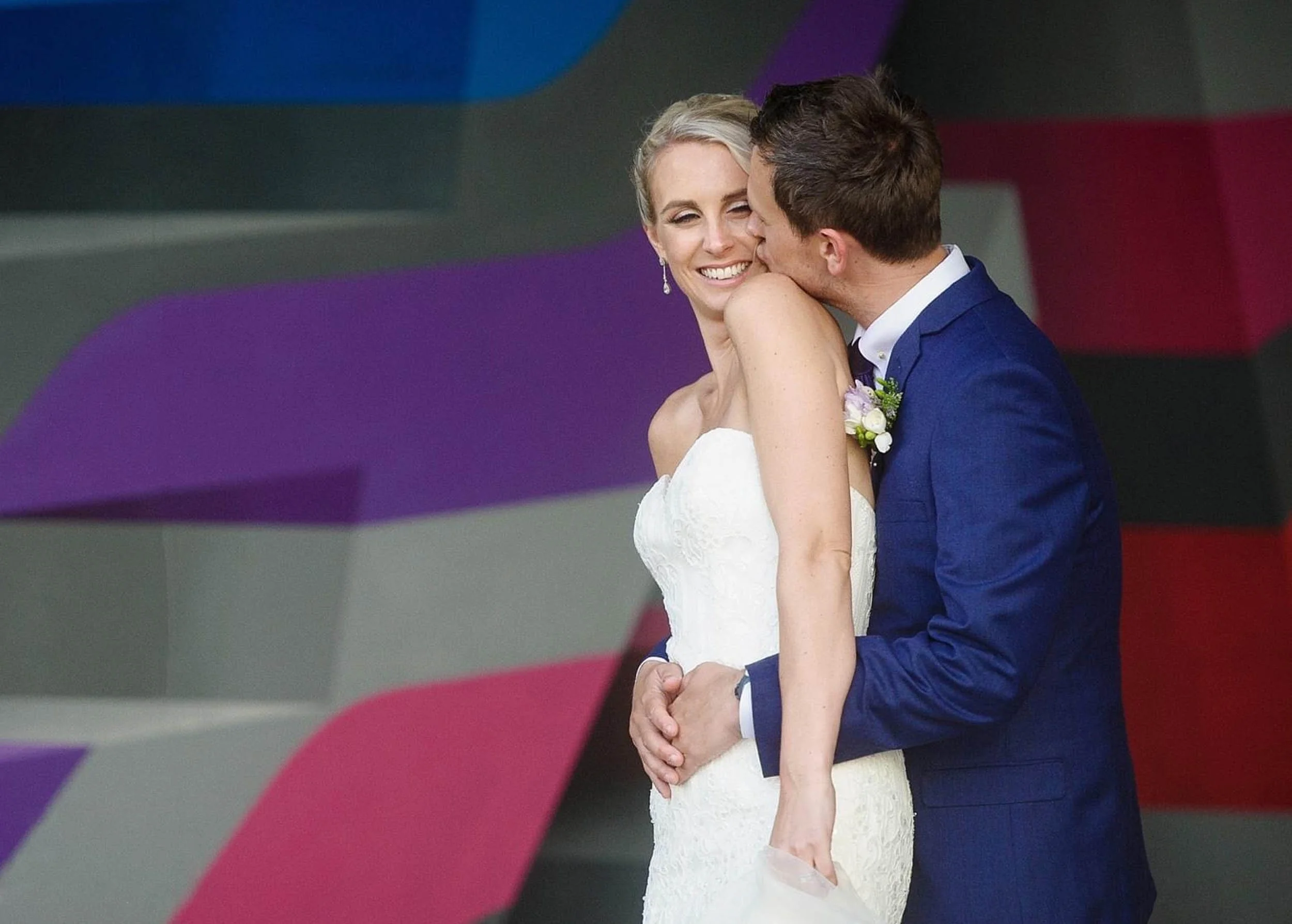 A bride and groom sharing a kiss, with the groom holding the bride's waist and the bride smiling in a strapless white wedding dress, in front of a colorful, abstract background.