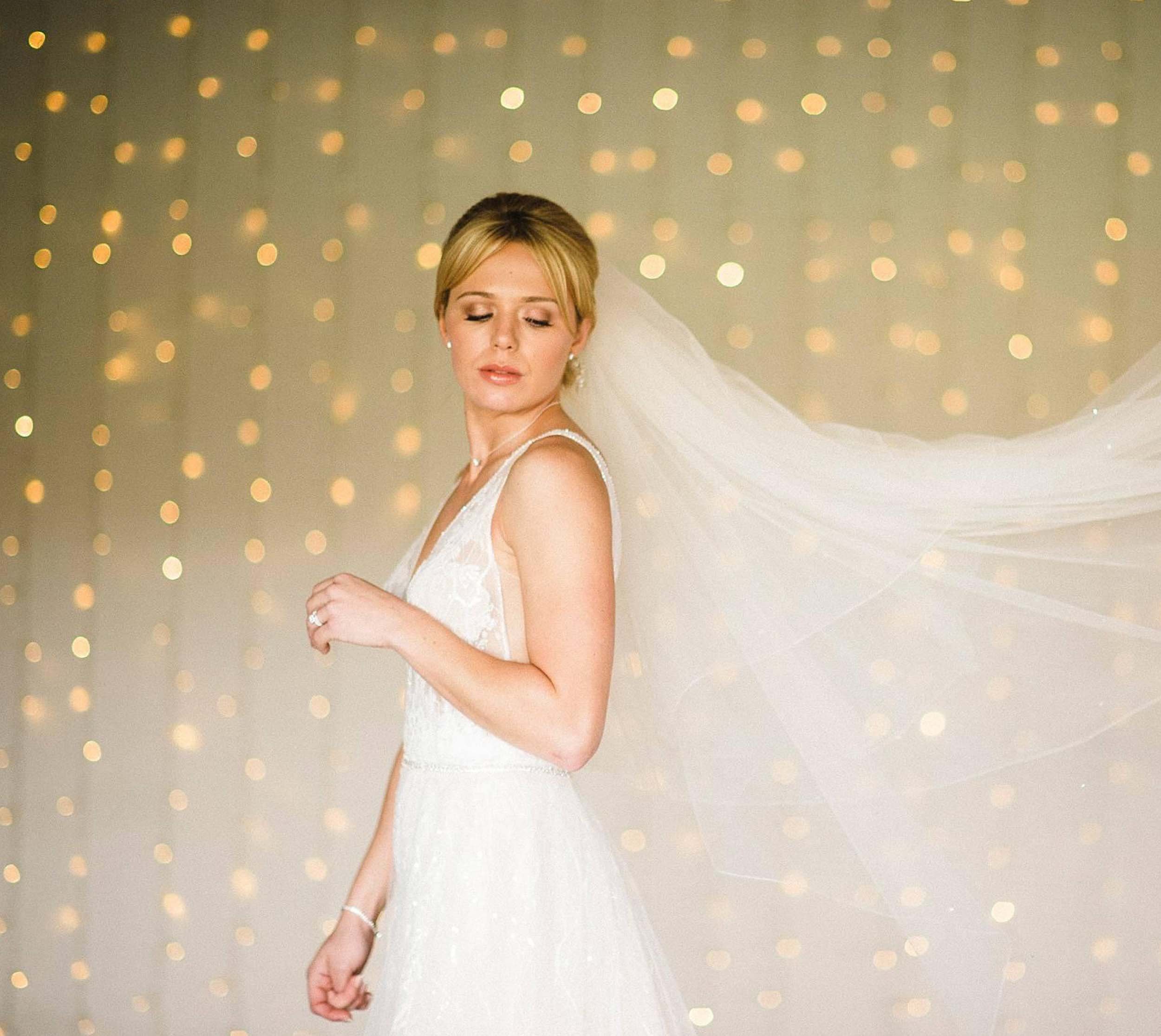 A bride in a white wedding dress with a veil, standing against a backdrop of blurred warm lights, with her eyes closed.
