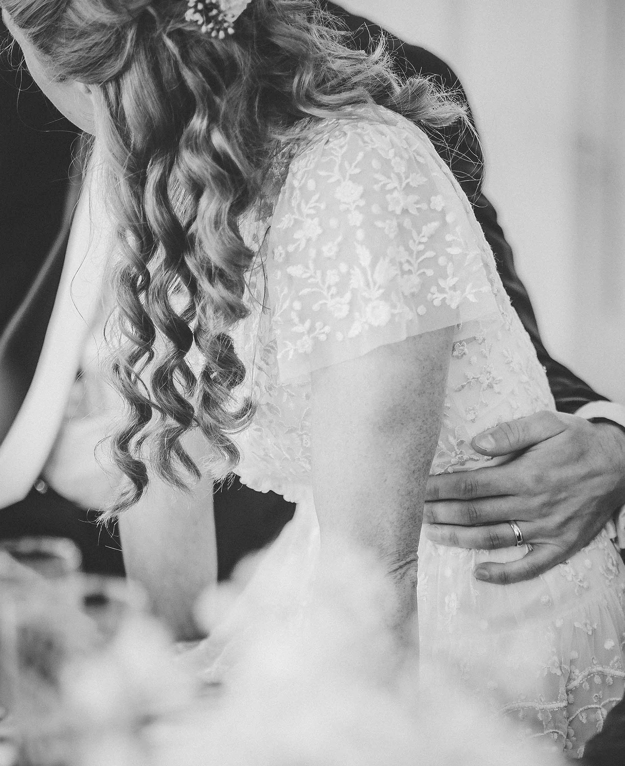 A bride with long, curly hair and a lace dress, with a hand gently placed on her waist.