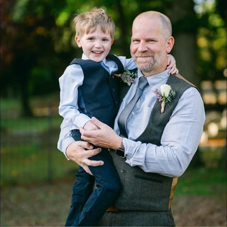 A man holding a young boy outdoors with trees in the background. The boy is smiling and wearing a formal vest and shirt. The man is wearing a light shirt and vest with boutonniere, indicating a special occasion.