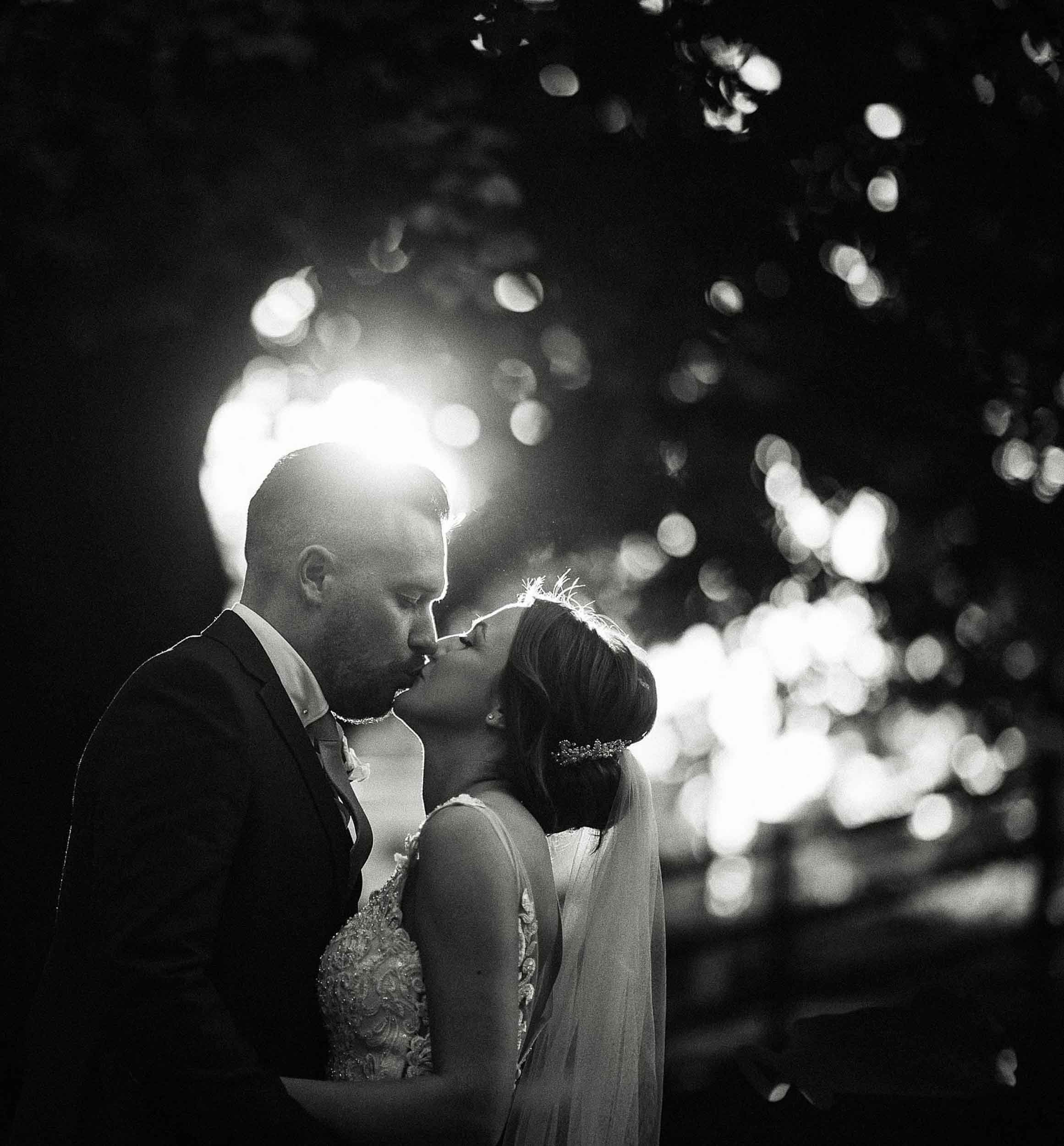 A black and white photo of a couple kissing, with the sun shining behind them and blurred trees in the background.
