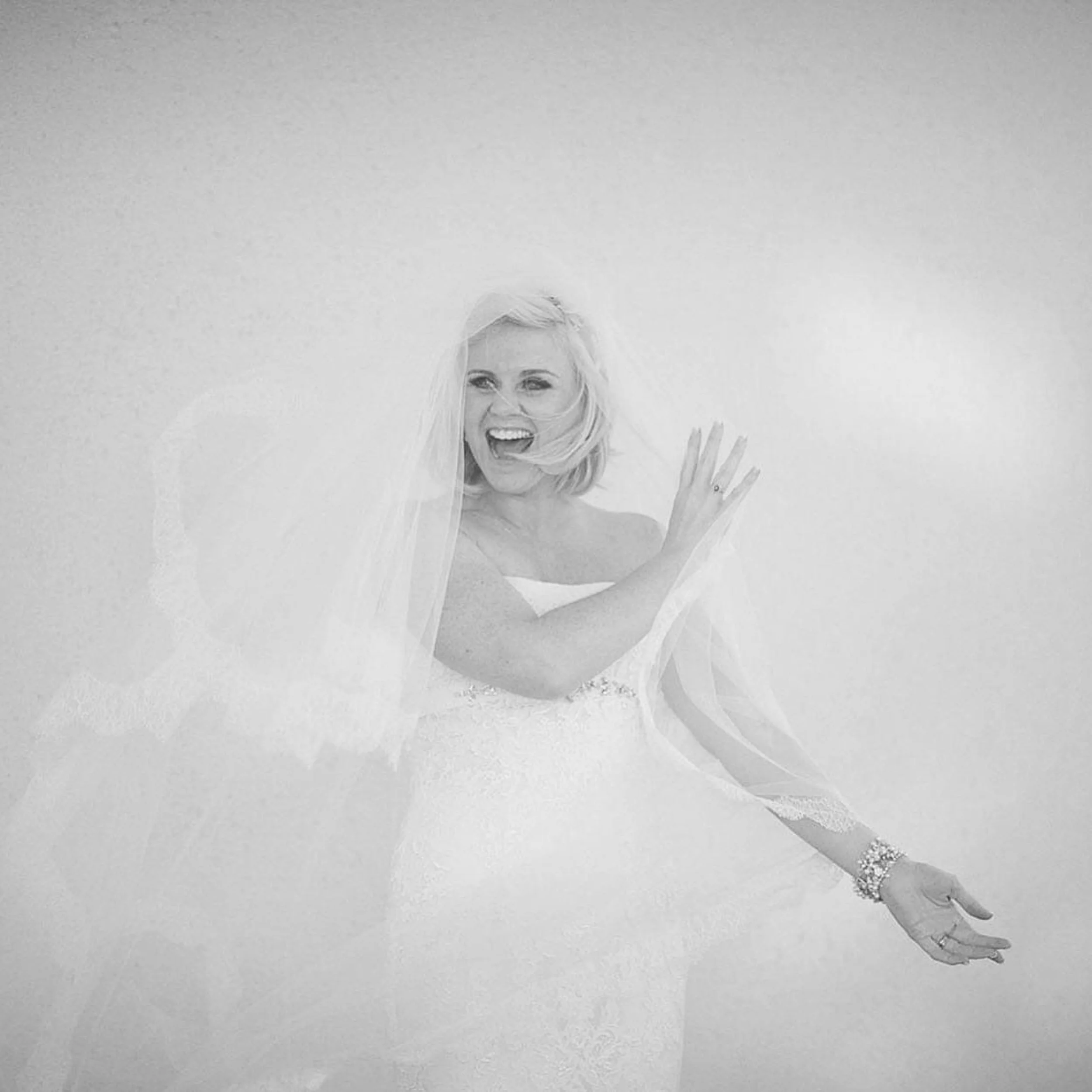 A woman with blonde hair in a wedding dress, smiling and waving with one hand while the other hand is outstretched, surrounded by a soft, glowing background.