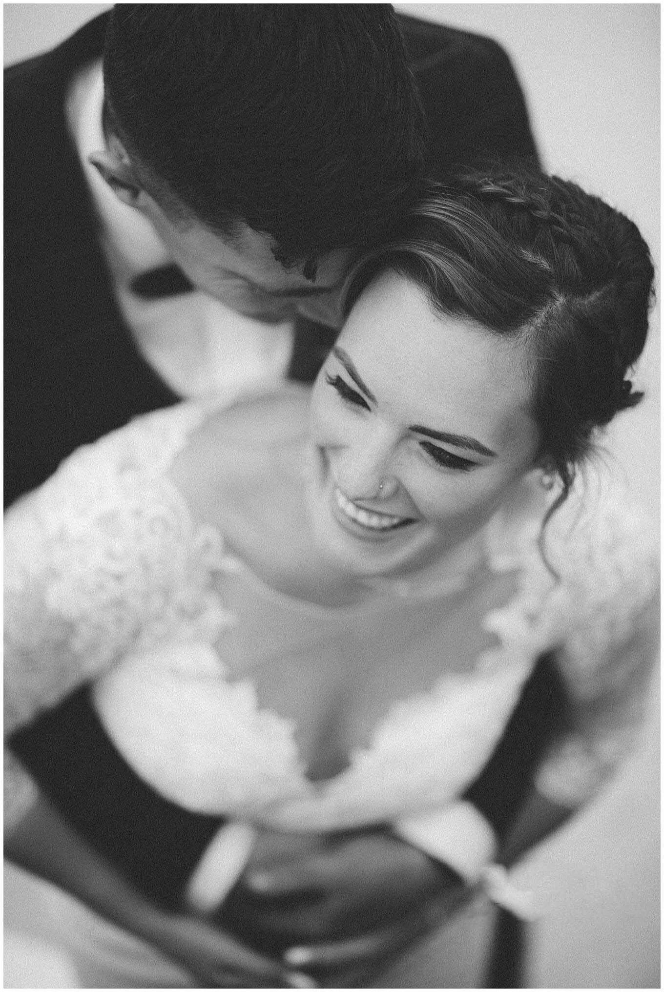 A black and white photo of a bride smiling with a groom kissing her on the forehead, with her in a wedding dress and the groom in a tuxedo.
