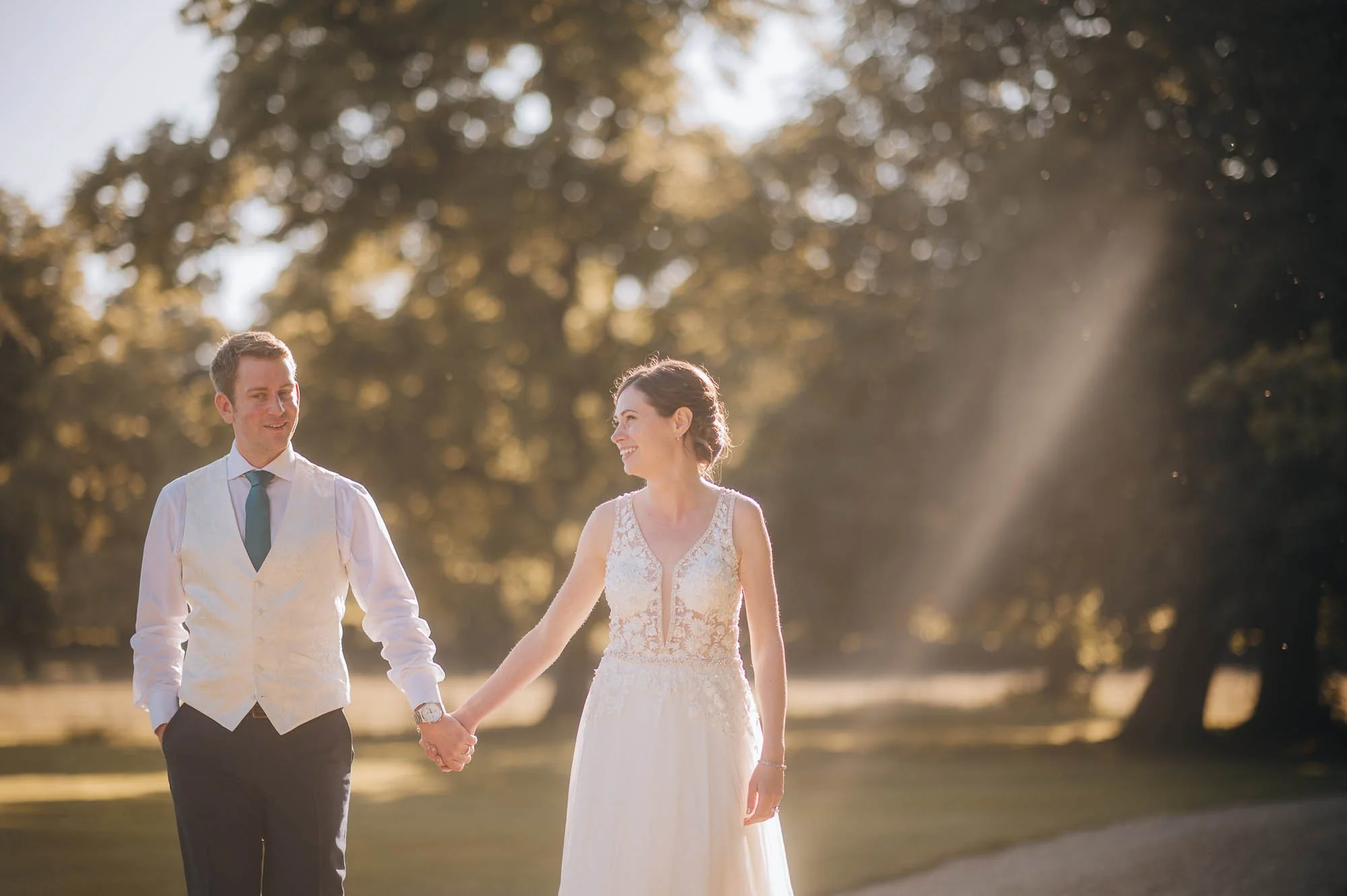 A bride and groom holding hands, walking outdoors on a sunny day, surrounded by trees.