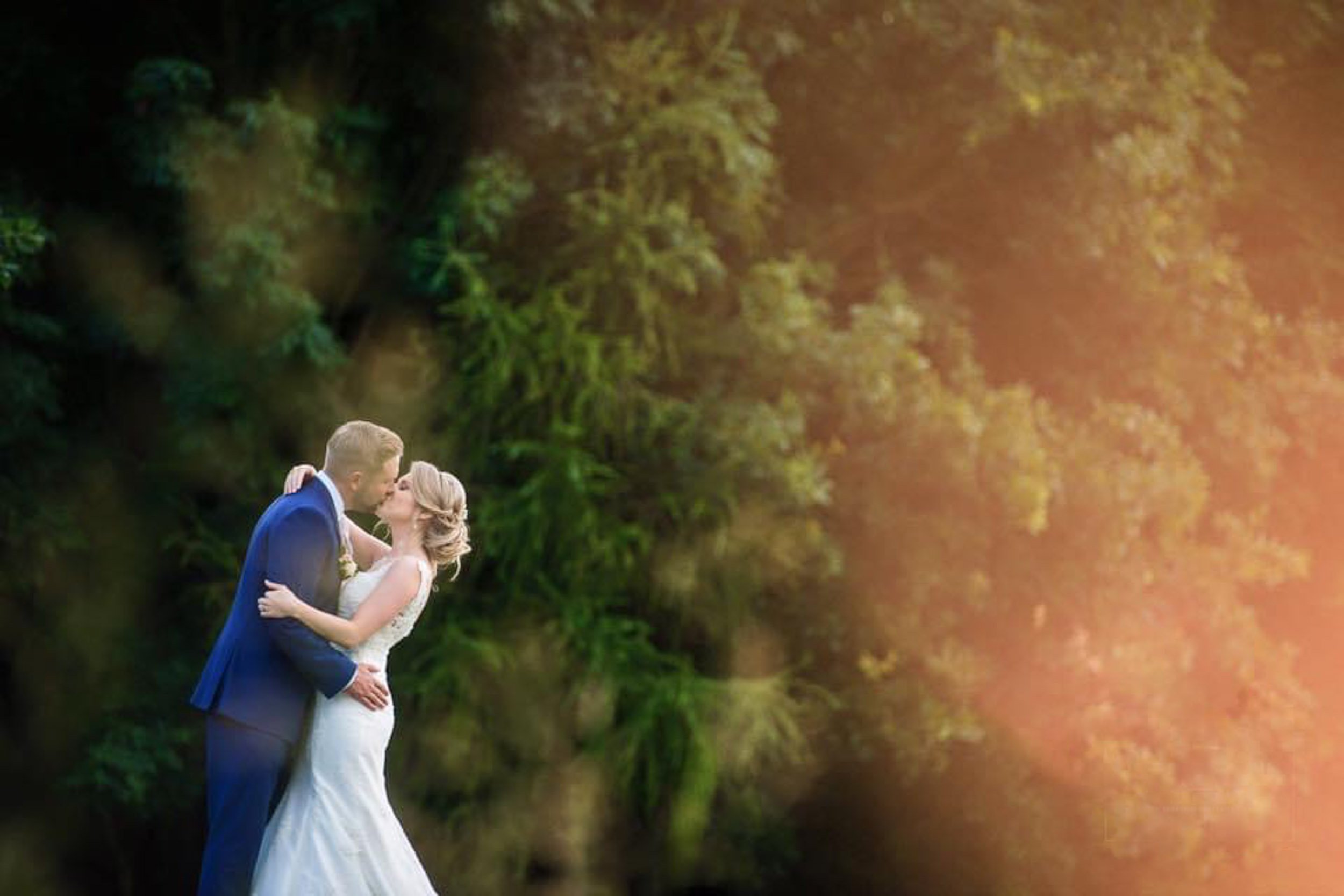 A newlywed couple sharing a romantic kiss outdoors in a forested area, with warm sunlight filtering through the trees.