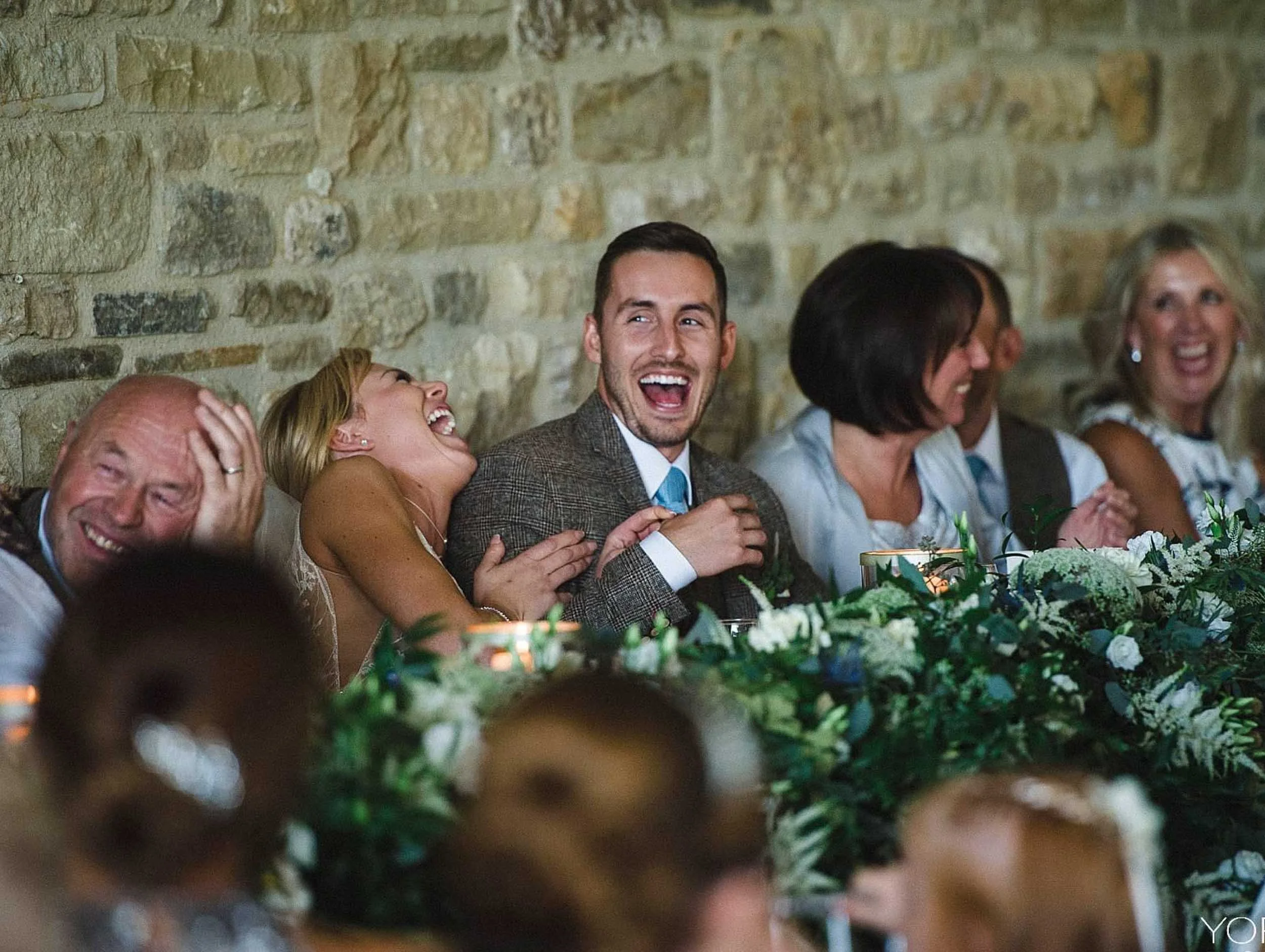 Group of people at a wedding reception, laughing and enjoying the moment, sitting against a stone wall with a floral centerpiece on the table in front of them.