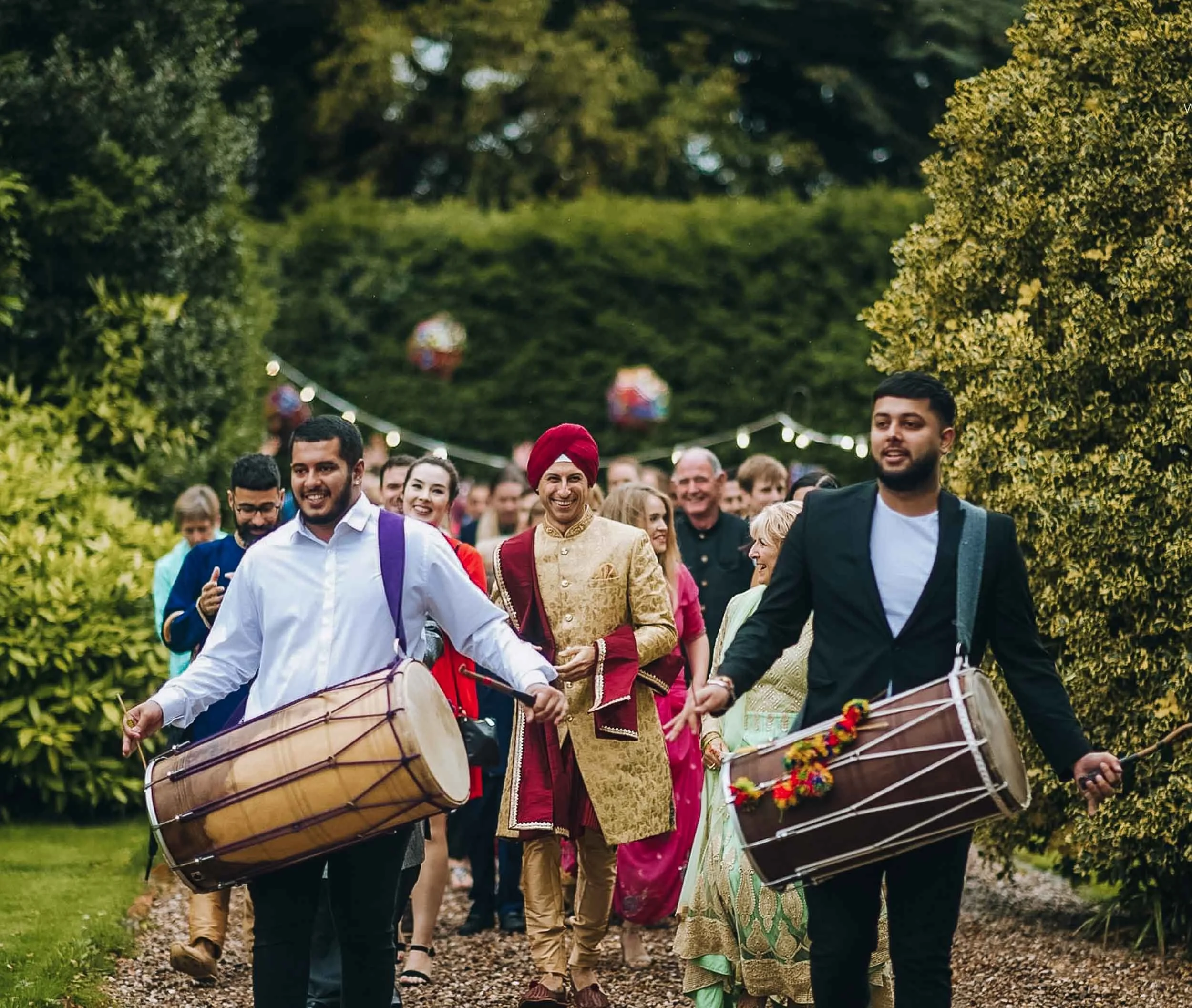 Group of people walking outdoors, some playing drums, in cultural attire, smiling, surrounded by greenery.