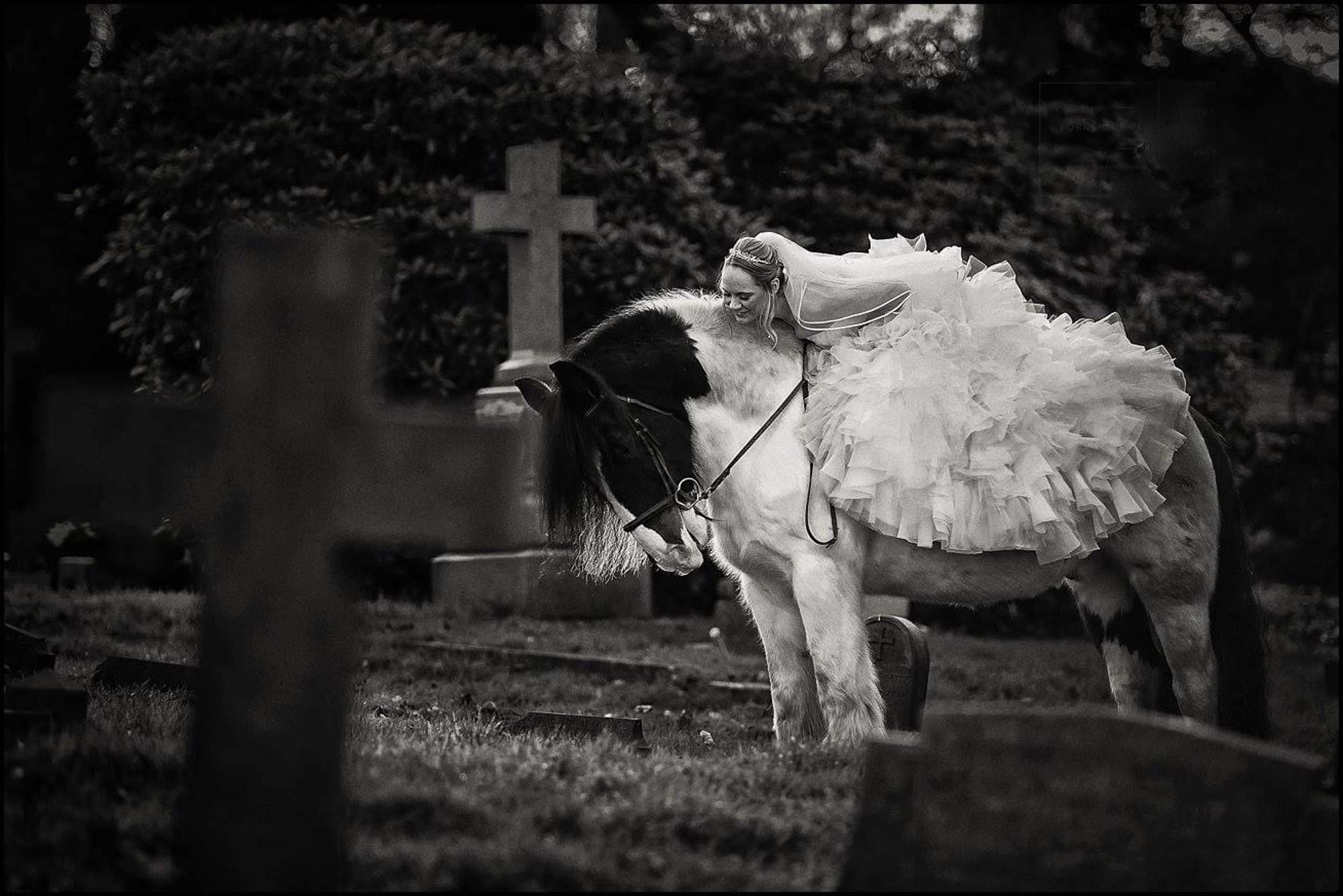 A woman in a wedding dress riding a horse in a cemetery, with gravestones and trees in the background.
