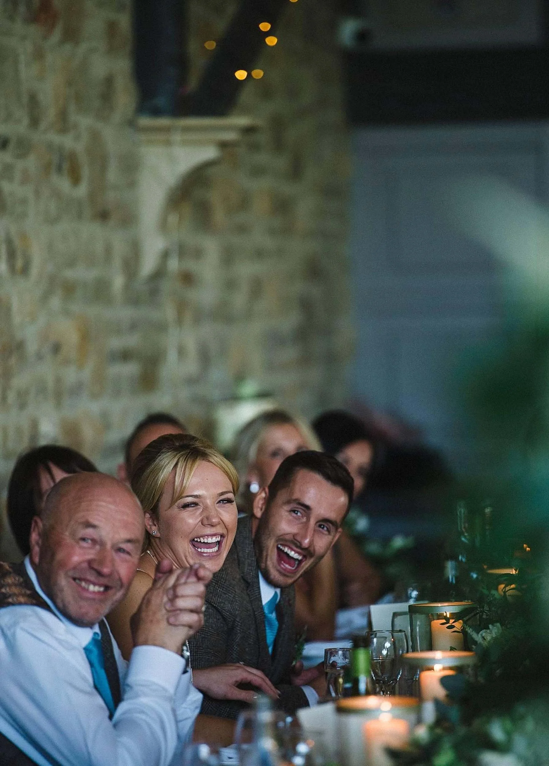 Group of smiling people sitting at a decorated dining table during a celebration or dinner event in a rustic setting, with candles and greenery on the table.